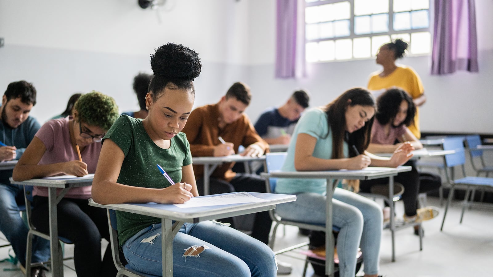 Several students write at desks in a classroom while a teacher walks by.