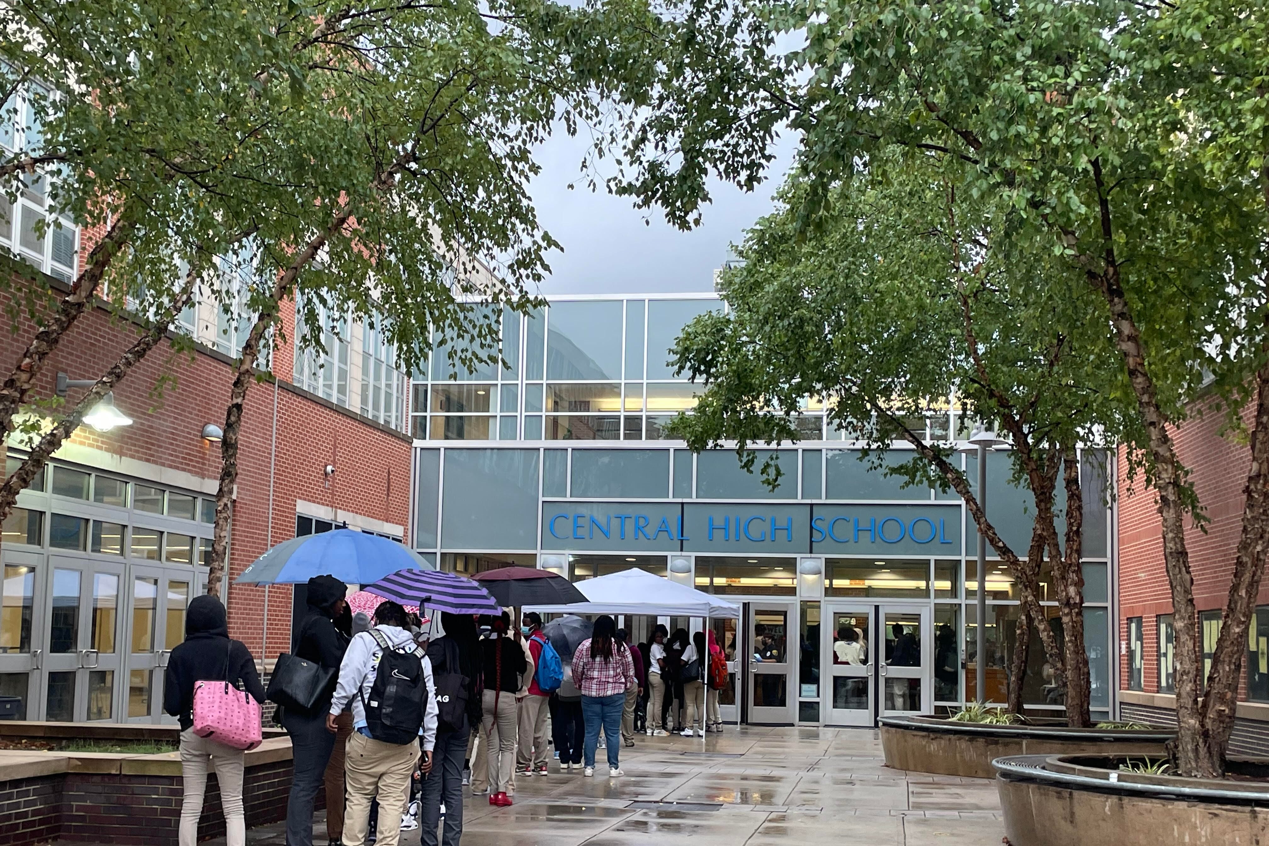 Students hold umbrellas as they stand in a line outside a high school with trees lining the entrance as it rains.