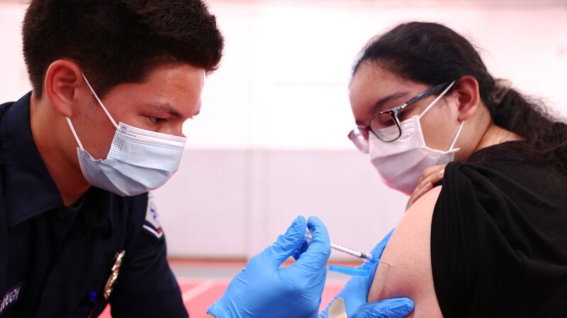 A masked and gloved health worker administers a vaccination in the arm of a masked patient.