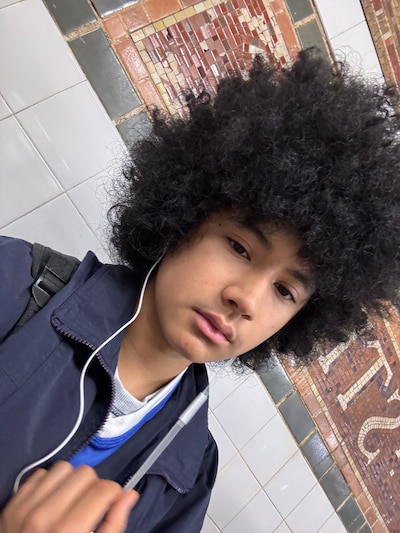 A photograph of a high school junior with a dark afro and wearing headphones poses for a selfie while walking down a sidewalk.