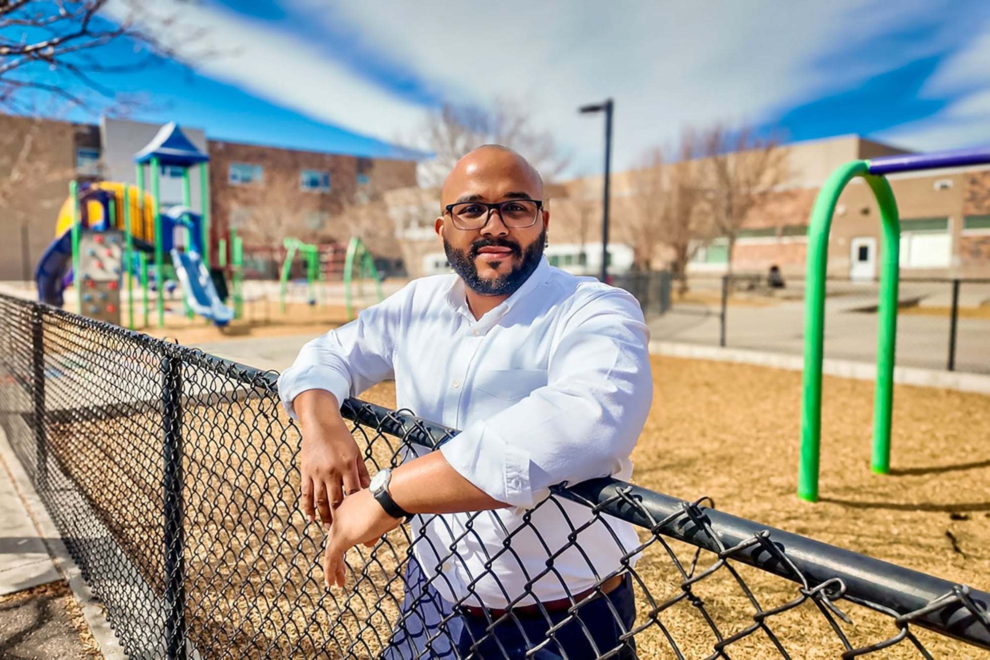 A bald man with a full beard and wearing a white dress shirt poses for a portrait while leaning on a school fence near a playground.