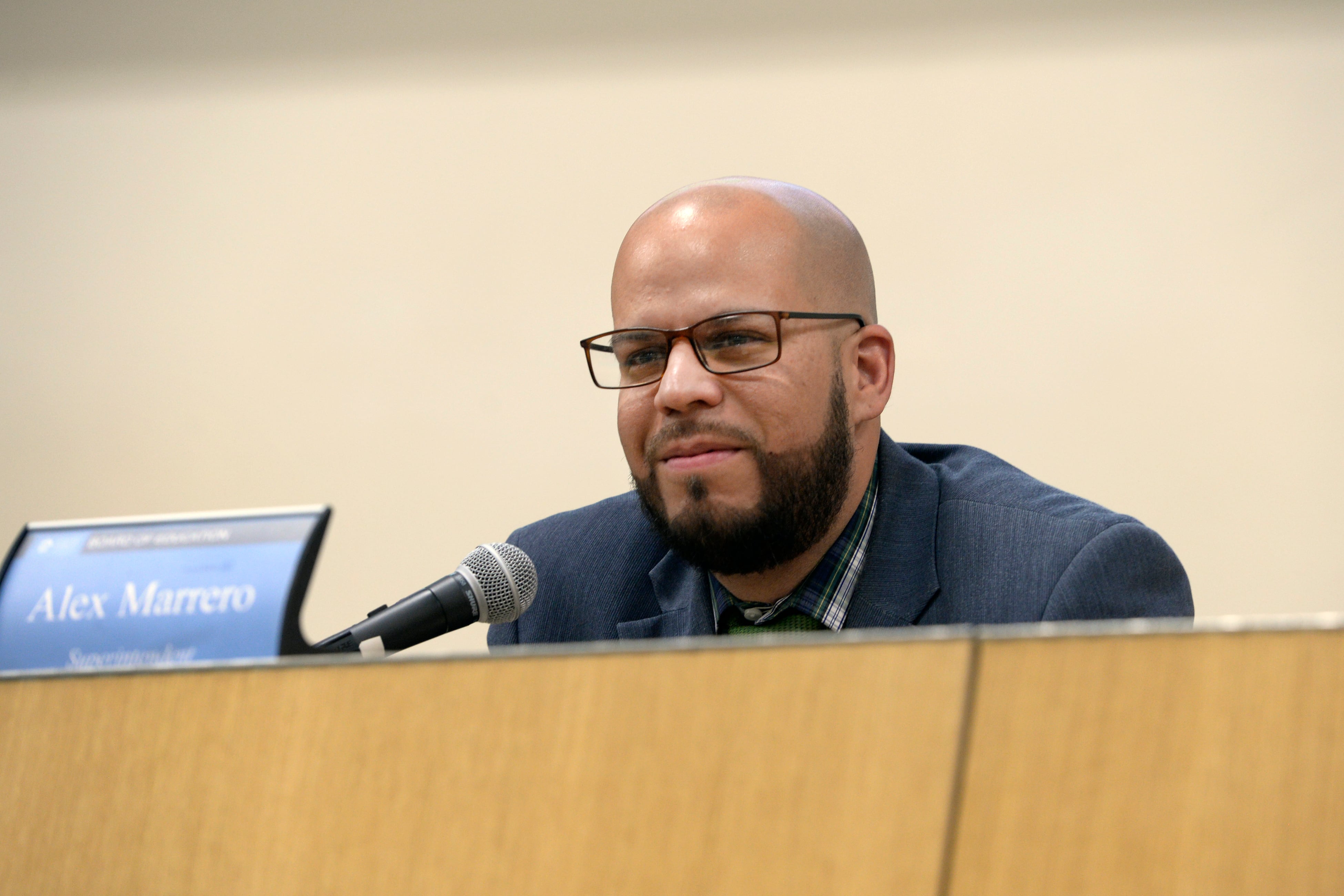 Denver Public Schools Superintendent sits on the dais with a microphone and nameplate in front of him at a school board meeting.