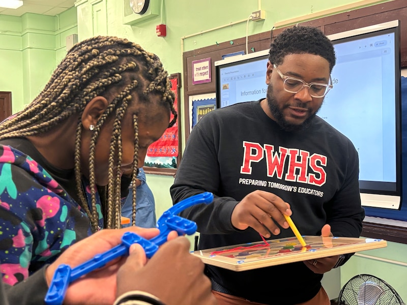 An adult in a black and red sweatshirt works on a board next to a student in a classroom.