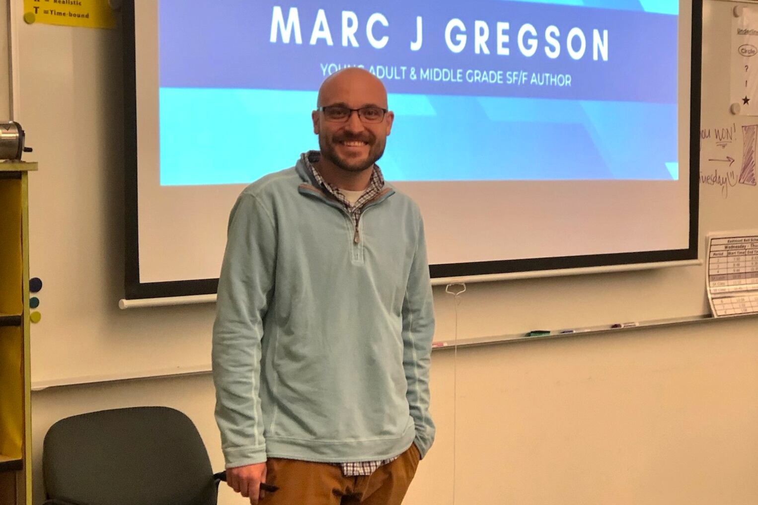 A man wearing a light blue sweater stands in front of a screen that says Marc J. Gregson.