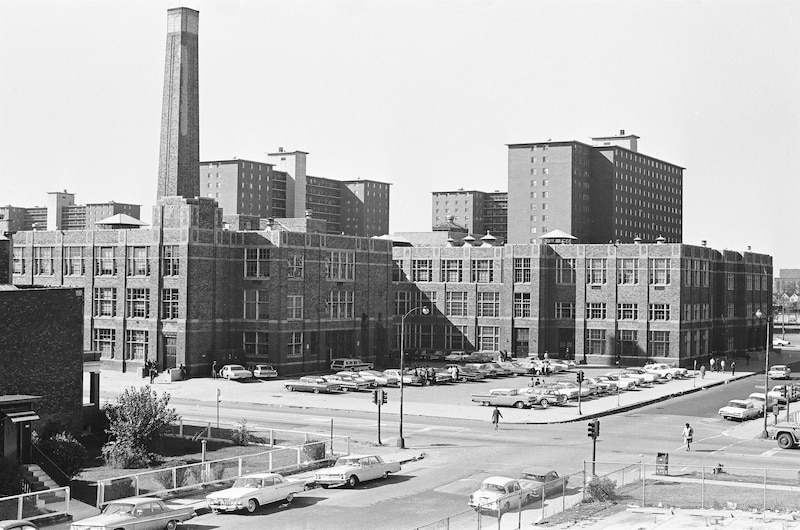A black and white photograph from 1966 with large three-story school building, with high rises looming behind it, and parked cars along a road and in a parking lot.