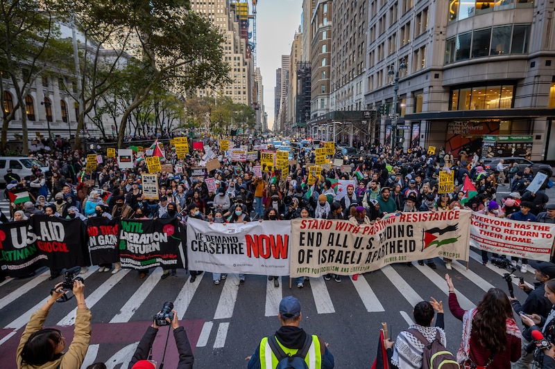 A mass group or protestors including students, educators and others fill a city street in NYC with a row of banners with various sayings supporting Palestine.
