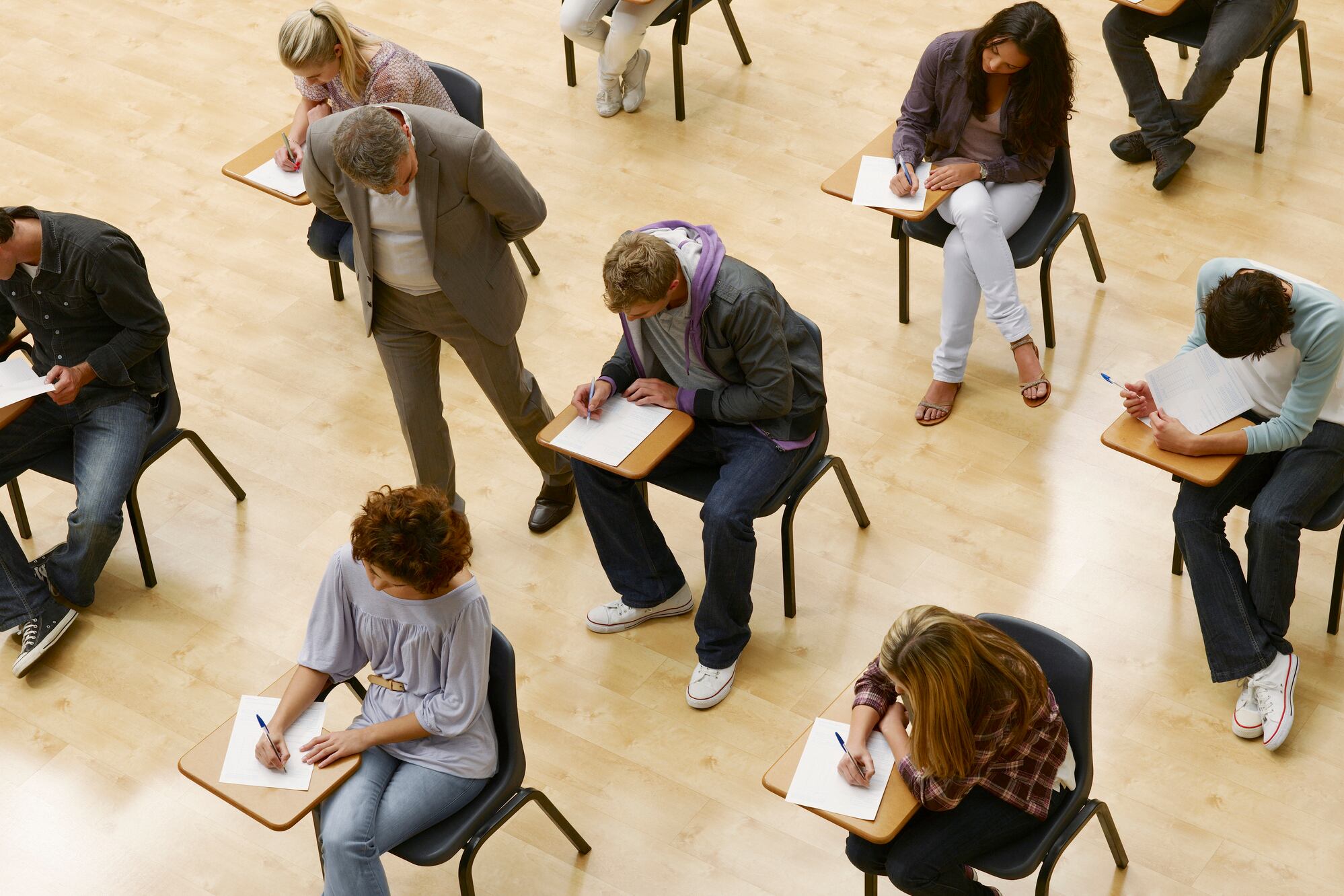 A group of students seated at desks take a test in a classroom with a teacher walking by.