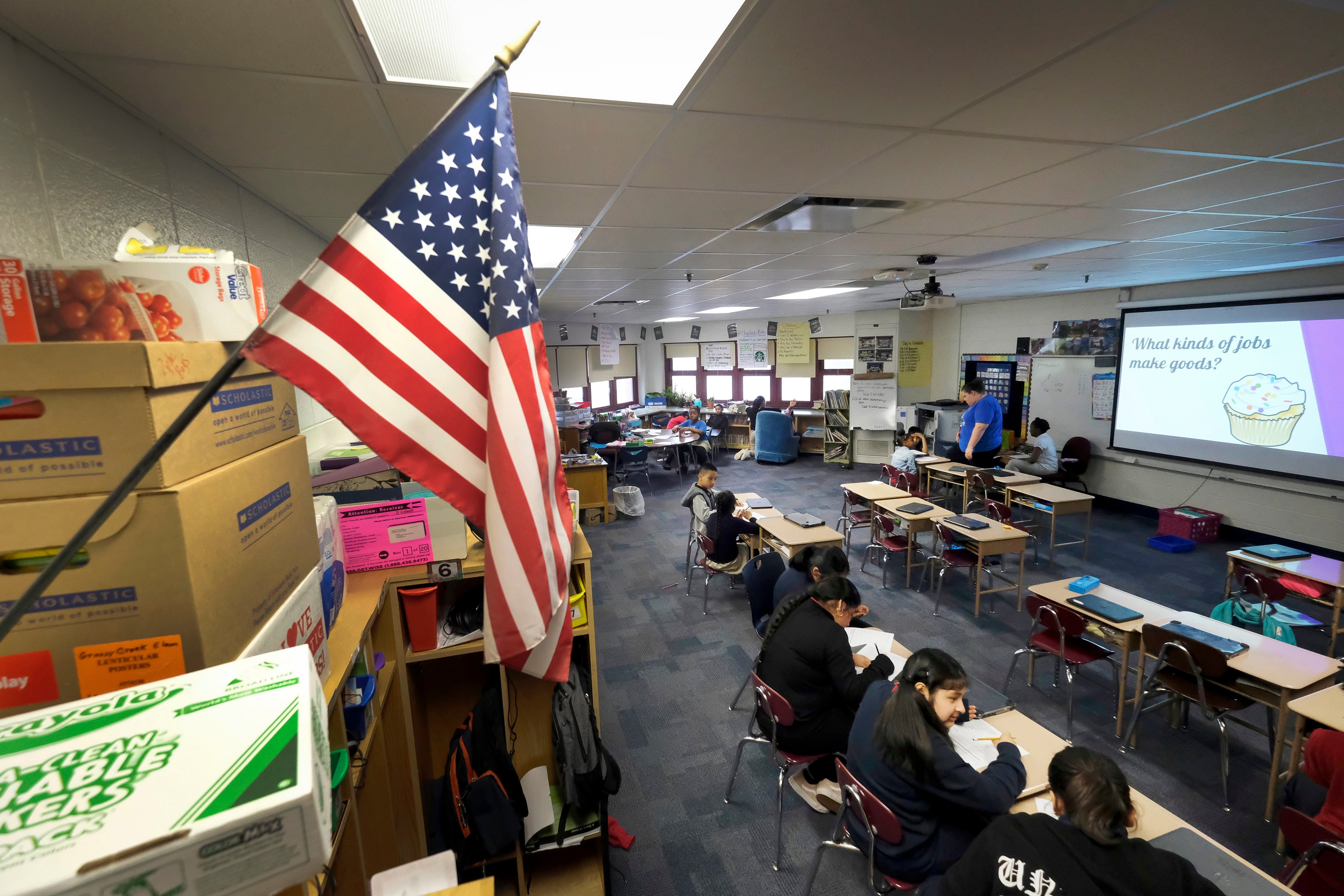 An American flag hangs on a wall an elementary classroom with a few students sitting in a row.