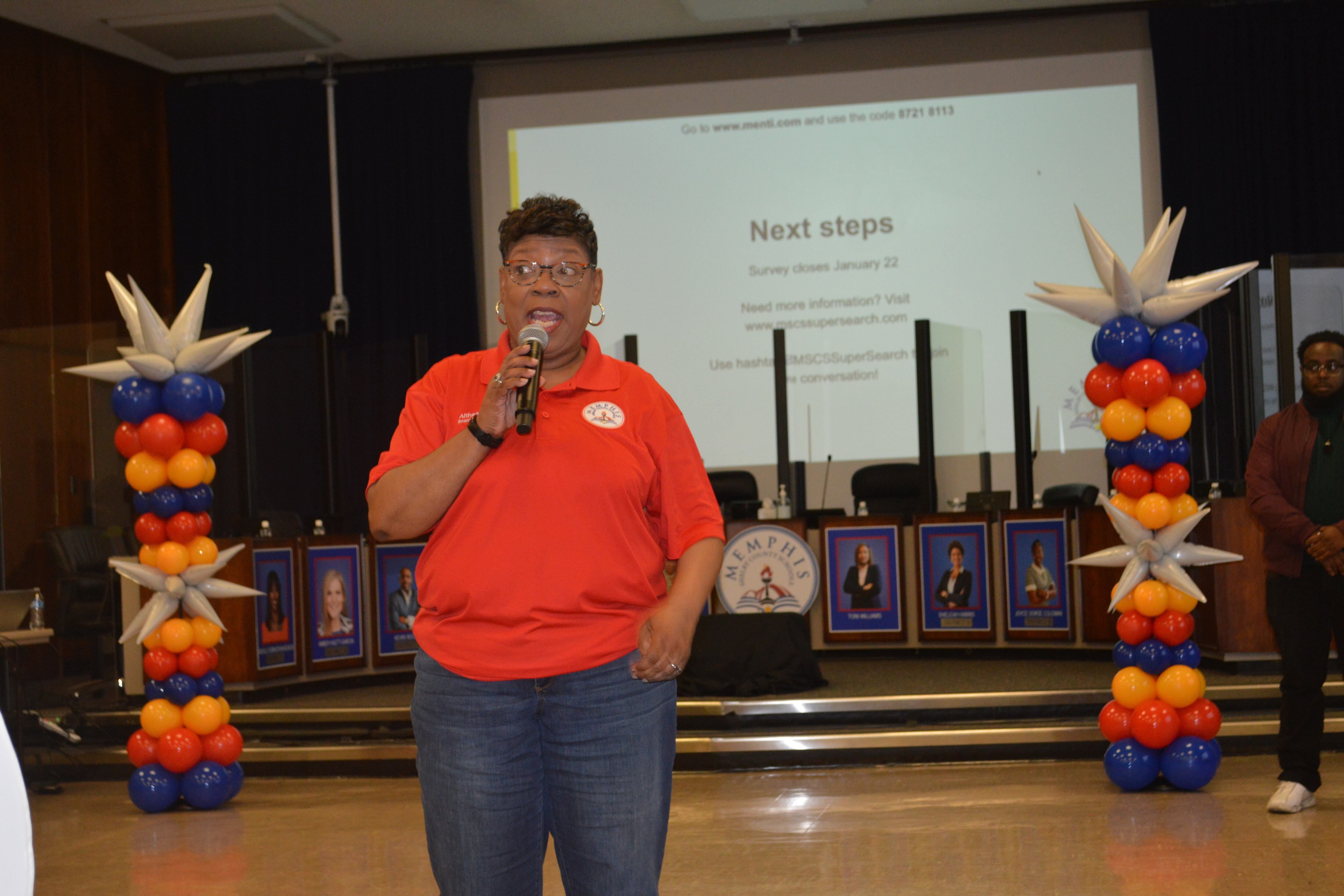 Woman in an red shirt and jeans holding a microphone, speaking in front of a stage.