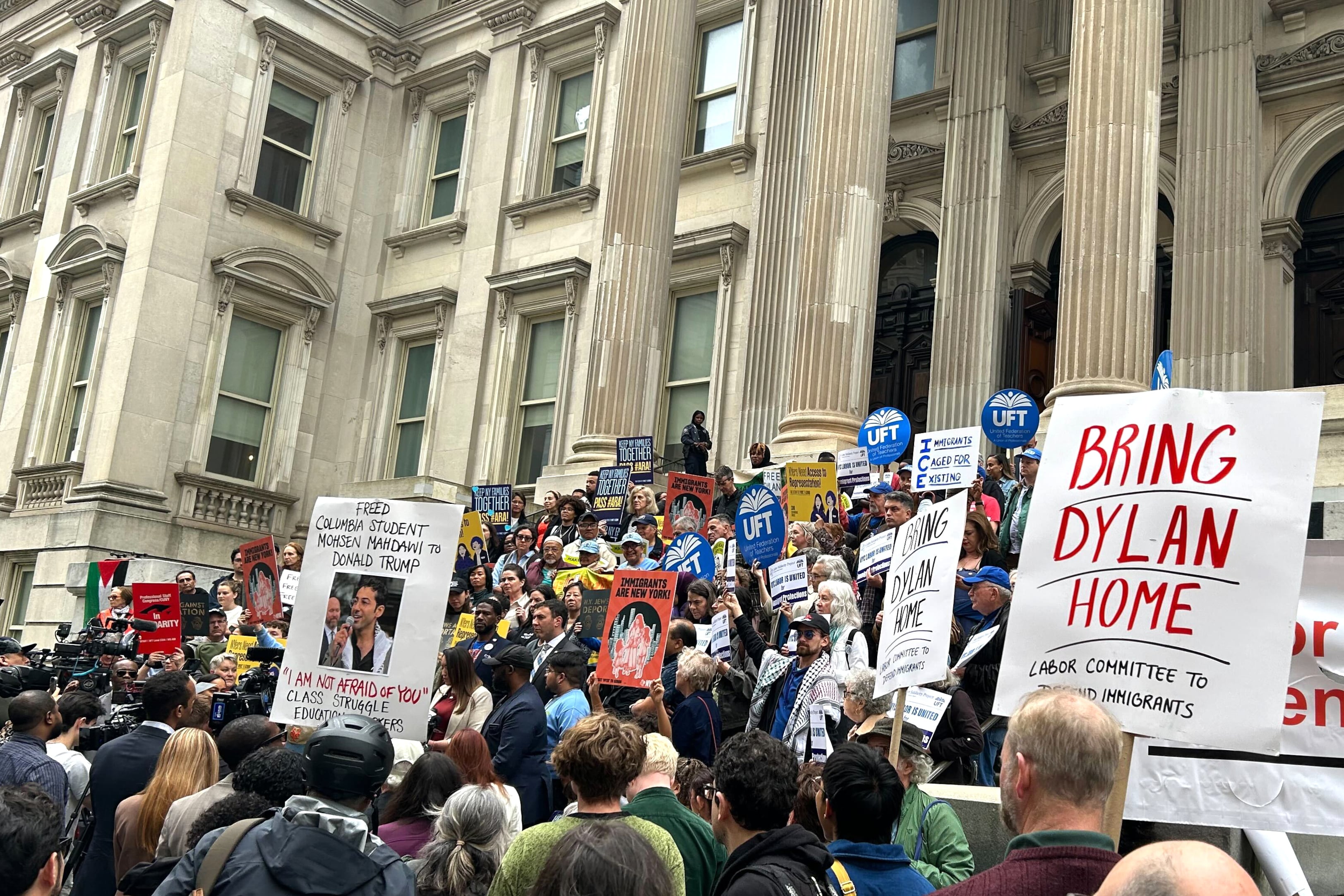 A crowd of people, some holding signs, stand on the steps of a building.