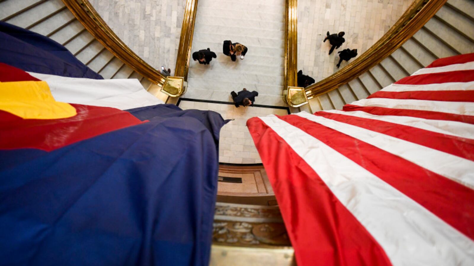 A Colorado state flag hangs next to a United States flag high above an entry way as people walk down a hallway.