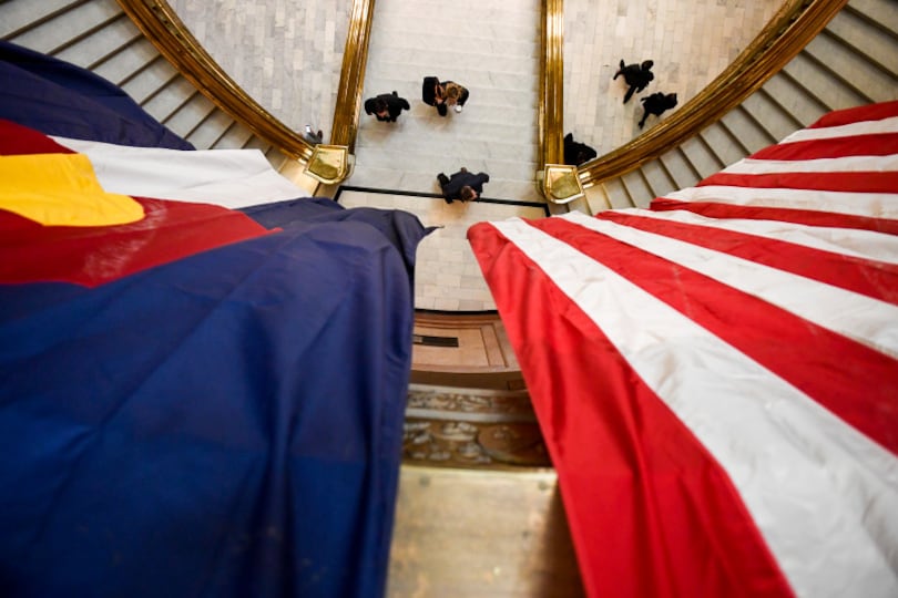 A Colorado state flag hangs next to a United States flag high above an entry way as people walk down a hallway.