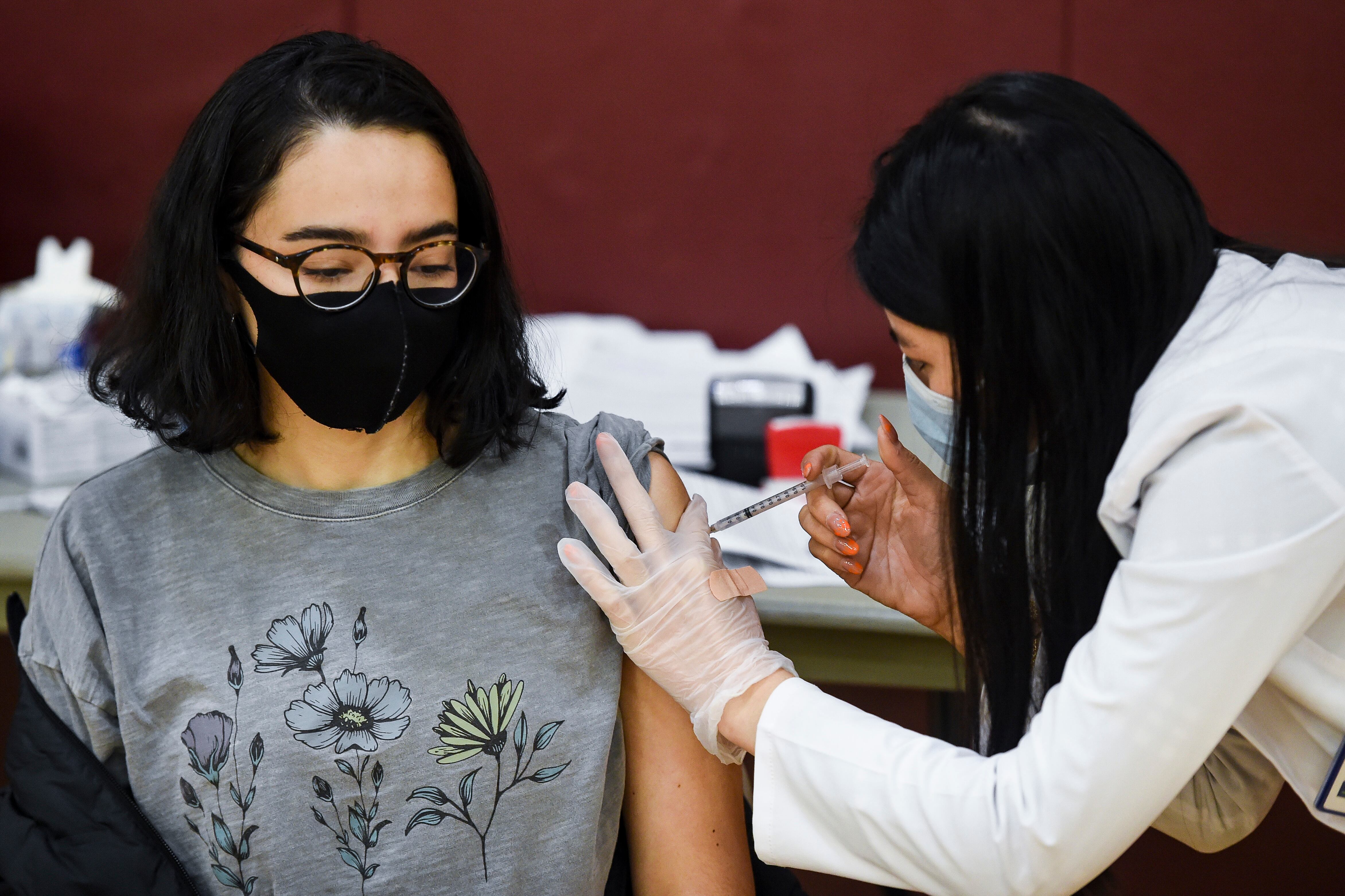 A young woman wearing glasses, a black mask and a grey t-shirt with flowers receives a vaccine from a woman wearing a blue surgical mask, gloves and white coat.