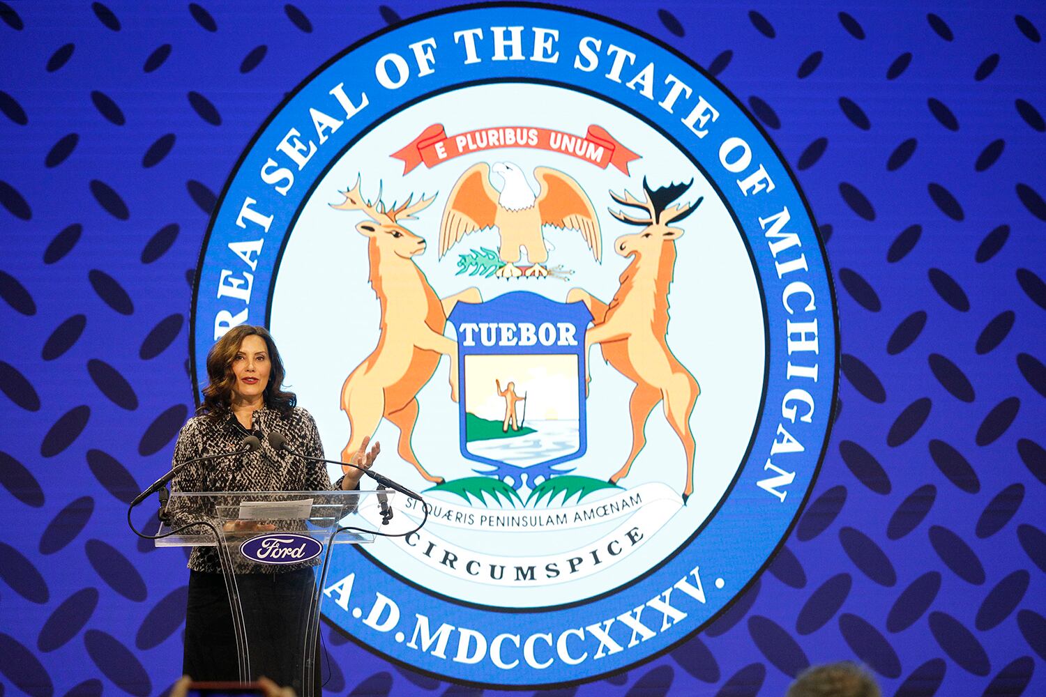 A woman wearing a dark top and a black skirt stands at a podium in front of a large Michigan state seal.
