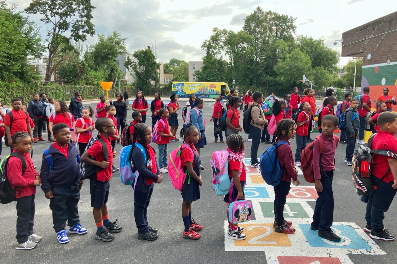 A photograph of a large group of elementary students wearing backpacks and some wearing red shirts while adults cheer them on.