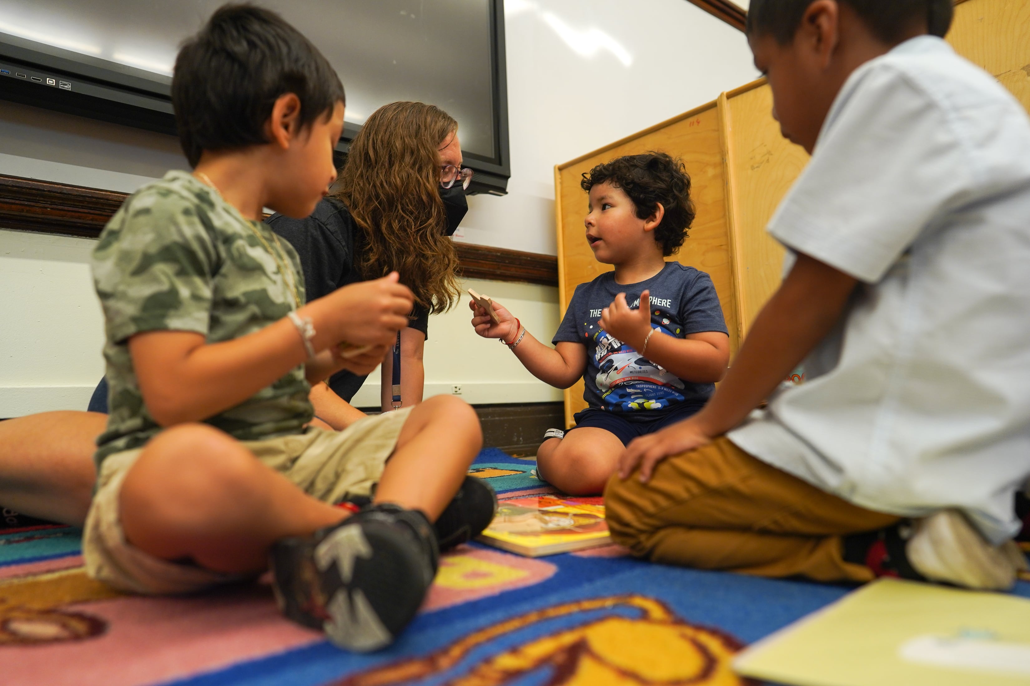 A woman teacher works with three young children on a multicolored rug in the classroom.