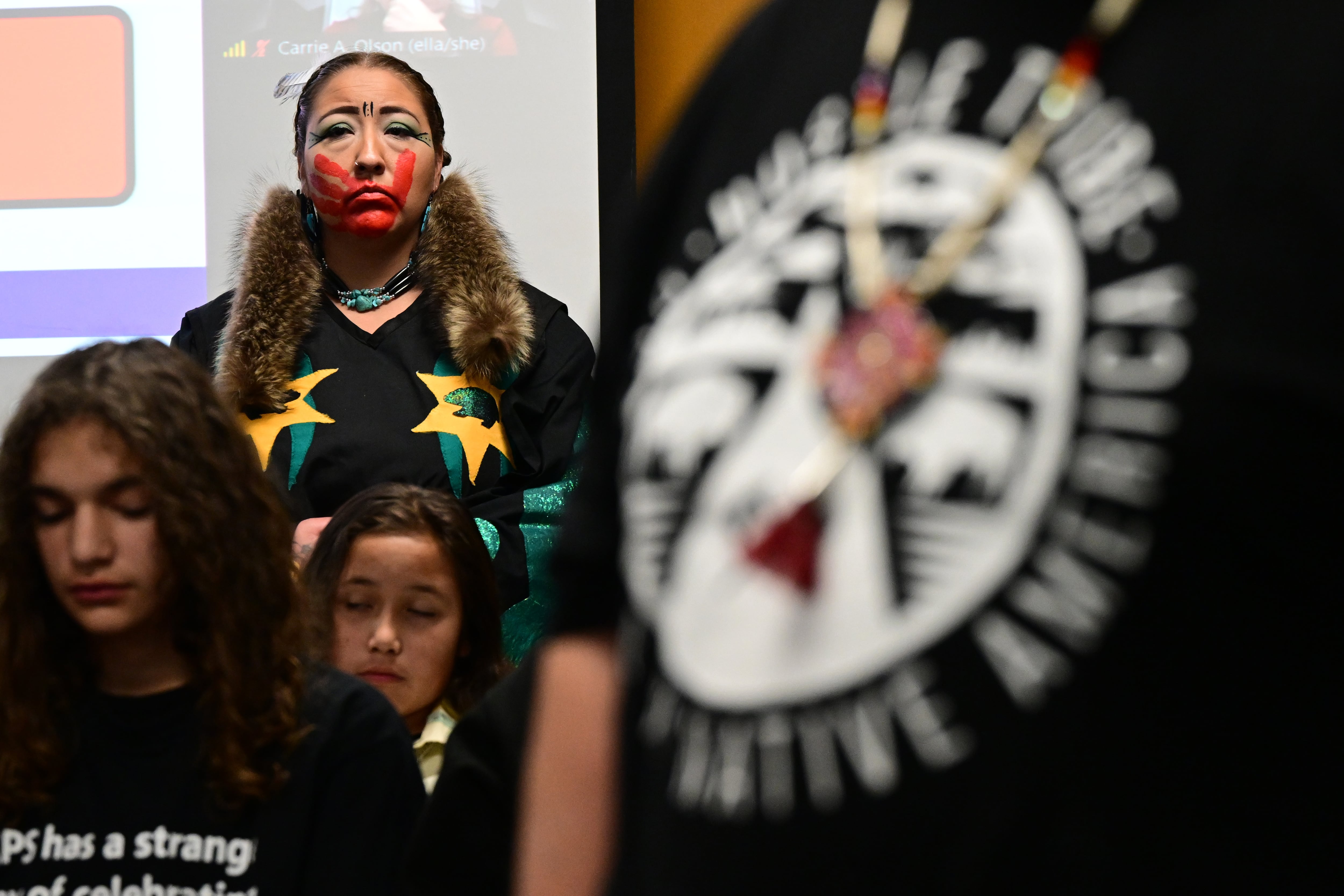 An Indigenous woman stands in the back of a crowded room listening to other speakers. She looks straight ahead. She has a large red handprint across her face. She is wearing a dark dress with yellow and blue stars.