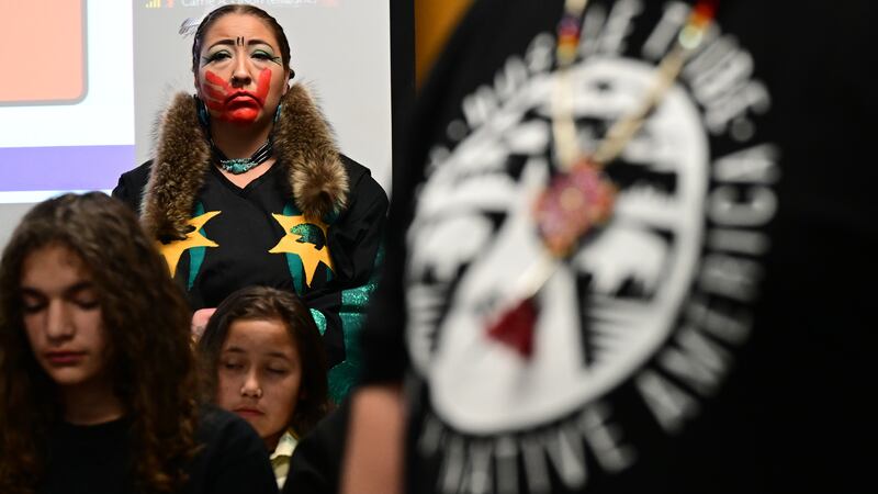 An Indigenous woman stands in the back of a crowded room listening to other speakers. She looks straight ahead. She has a large red handprint across her face. She is wearing a dark dress with yellow and blue stars.