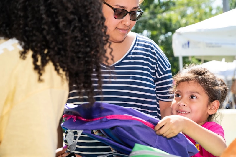 A little girl picks up a purple backpack while a woman in a striped shirt looks at her.