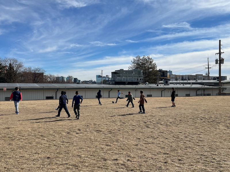 A group of young students play soccer outside with a blue sky and clouds in the background.