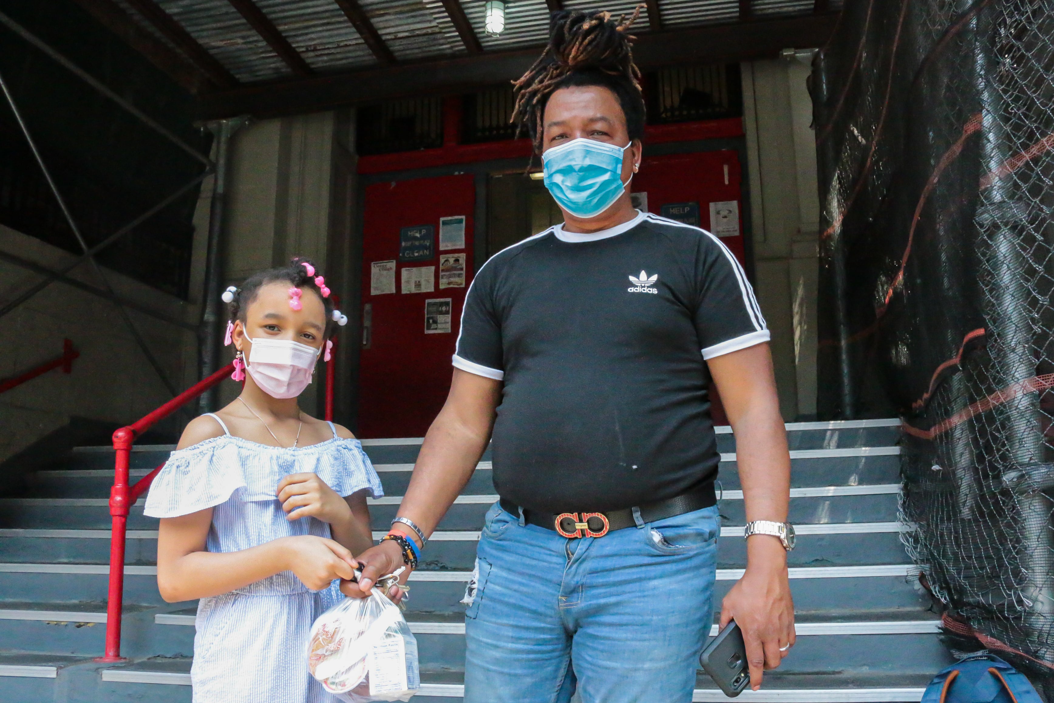 A young girl wearing a white dress, pink surgical mask, and pink and white jewelry in her hair holds her father’s hand, who is wearing a black Adidas shirt, blue jeans and a blue surgical mask.