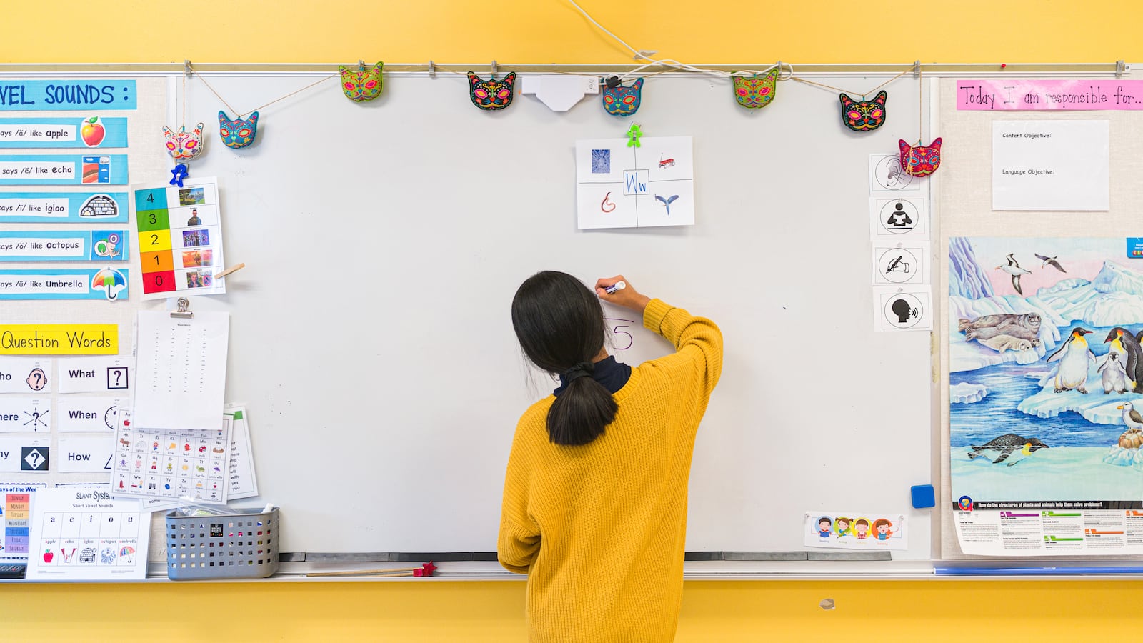 A student, wearing a yellow sweater, writes on a whiteboard that hangs on a bright yellow wall.