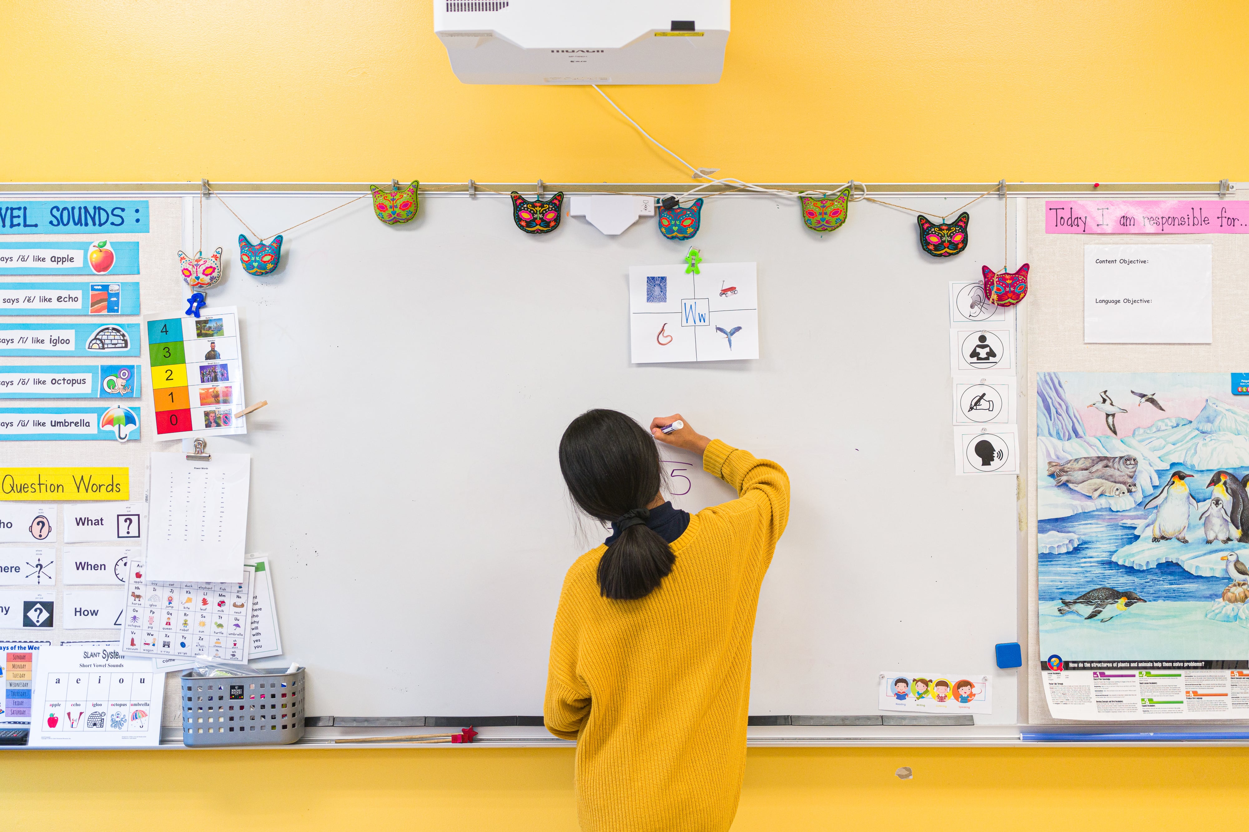 A student, wearing a yellow sweater, writes on a whiteboard that hangs on a bright yellow wall.