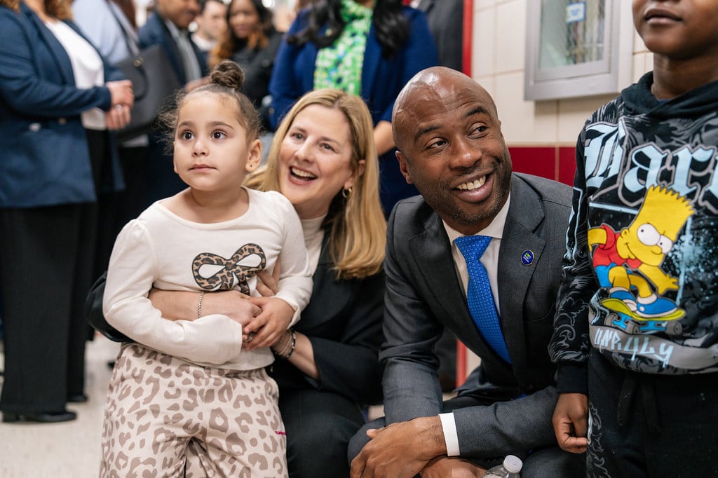 A photograph of a Black man in a suit kneeling next to a white woman and two young children in a school visit.