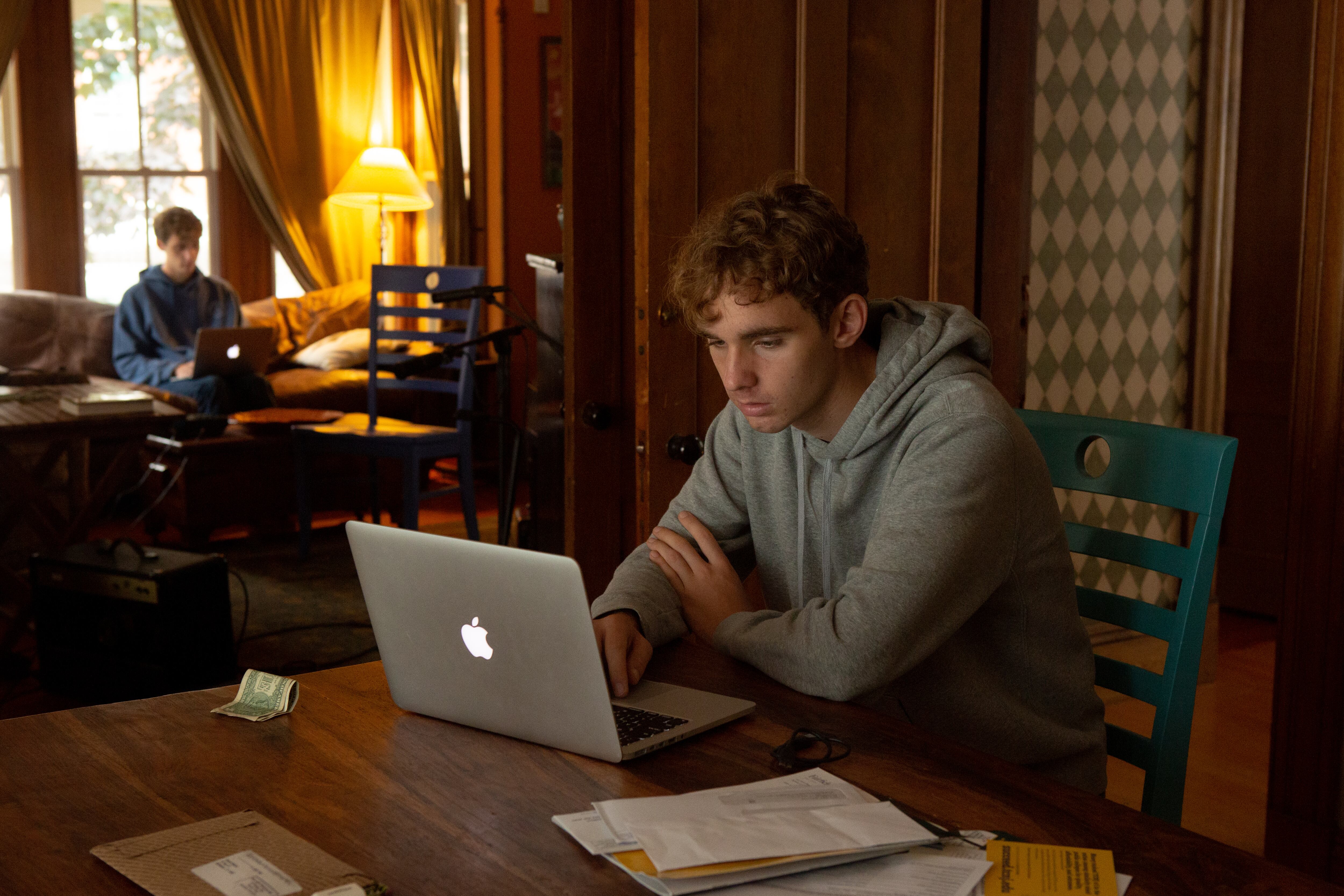 One teenage boy works on a laptop at a table at home, while another sits on a sofa against windows in the background.