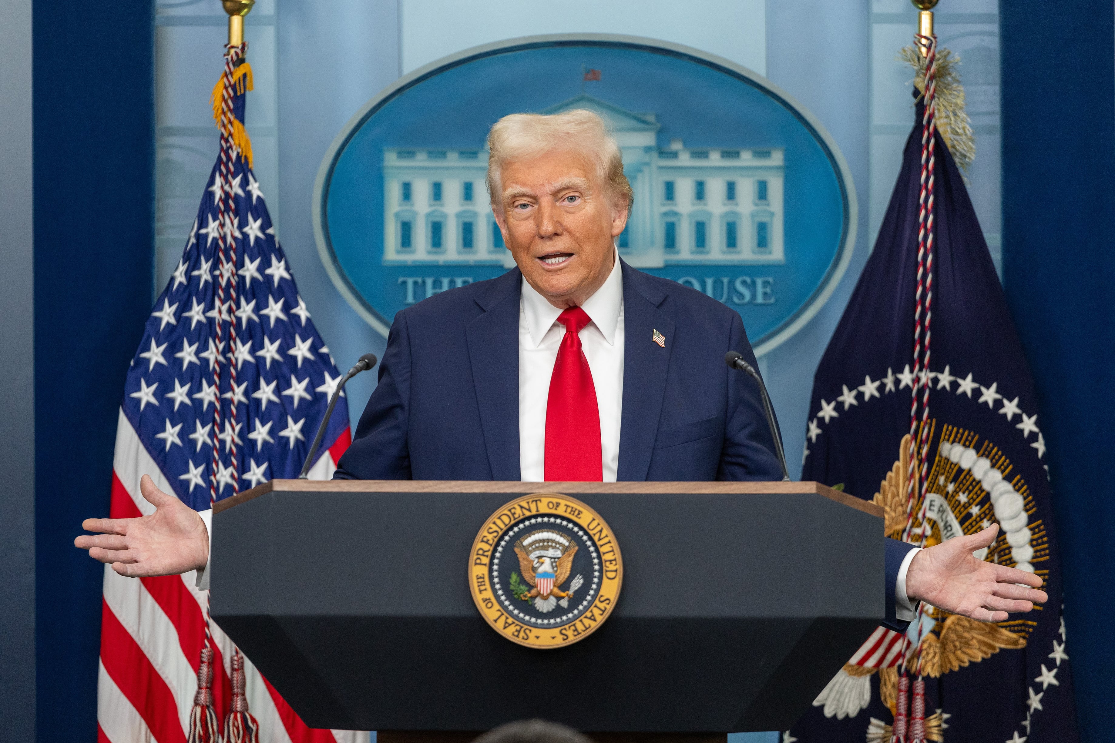 President Donald Trump in a dark blue suit and red tie stands behind a podium and in front of a background with two flags.