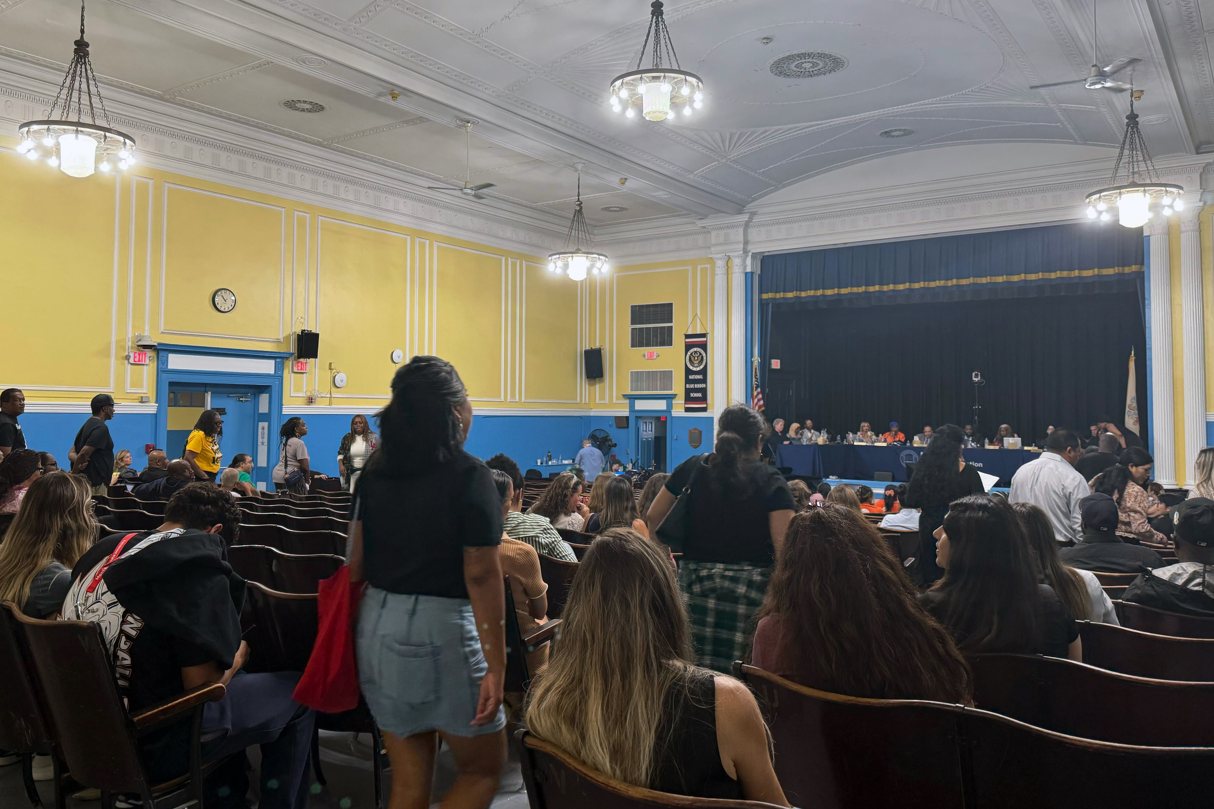 A photograph of a school auditorium filled with adults watching a row of adults sitting on stage.