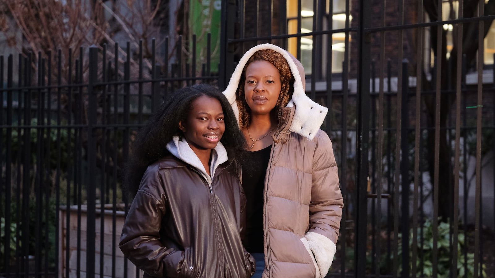 An adult woman and her daughter wear winter jackets and stand next to each other posing for a portrait outside with a black gate in the background.