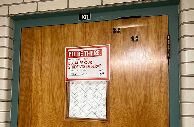 A wooden classroom door has a white and red sign on it.
