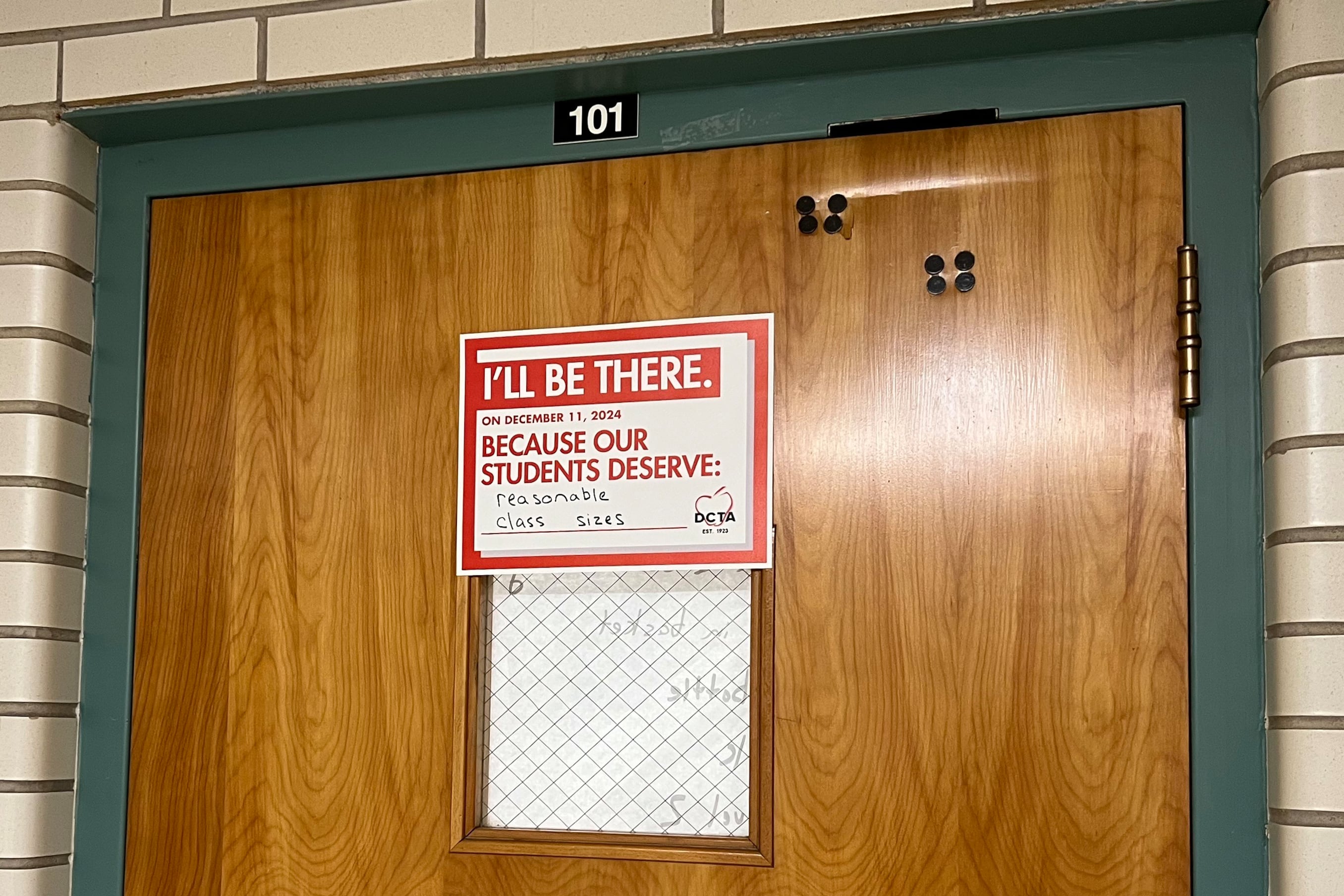 A wooden classroom door has a white and red sign on it.