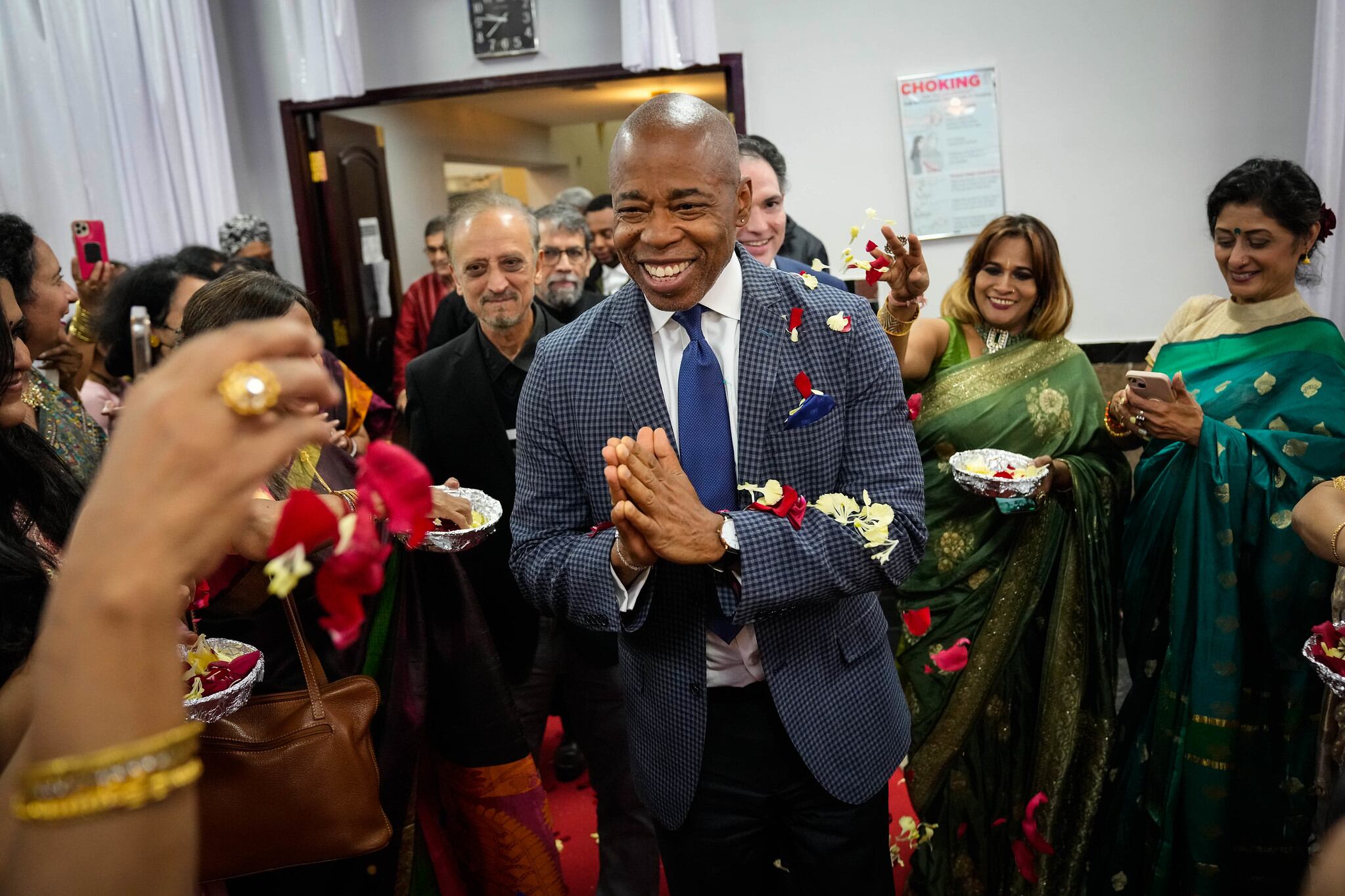 Mayor Eric Adams in a blue suit and tie stands in front of two women dressed in green saris.