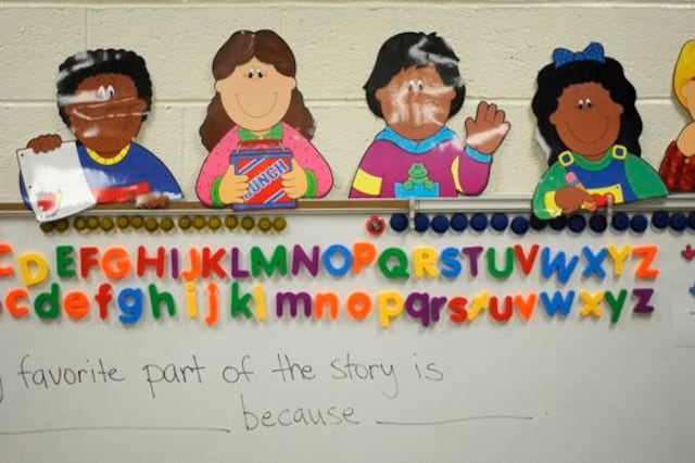 Cartoon cutouts of children are taped in a row on a wall with magnetic alphabet letters on a whiteboard.
