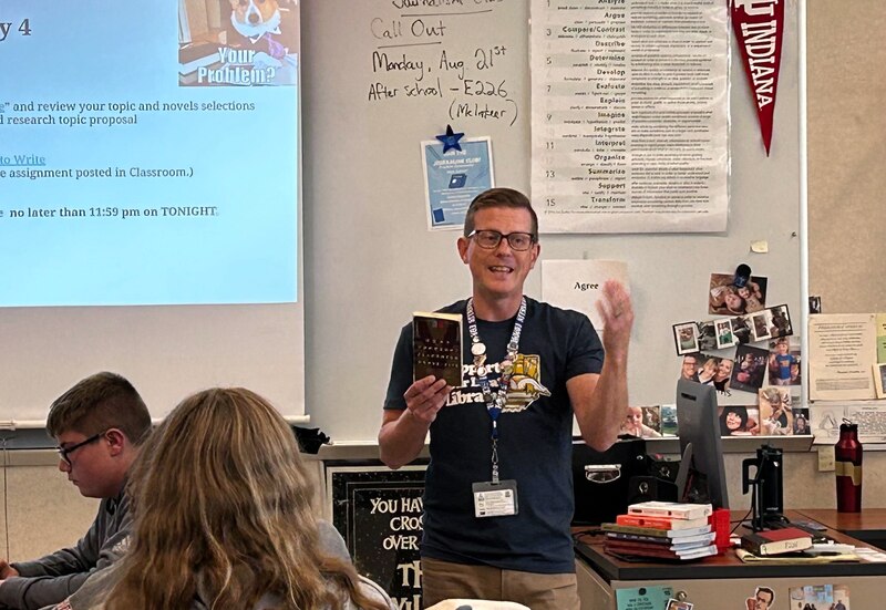 A man with glasses stands in front of a class with two students in sight.