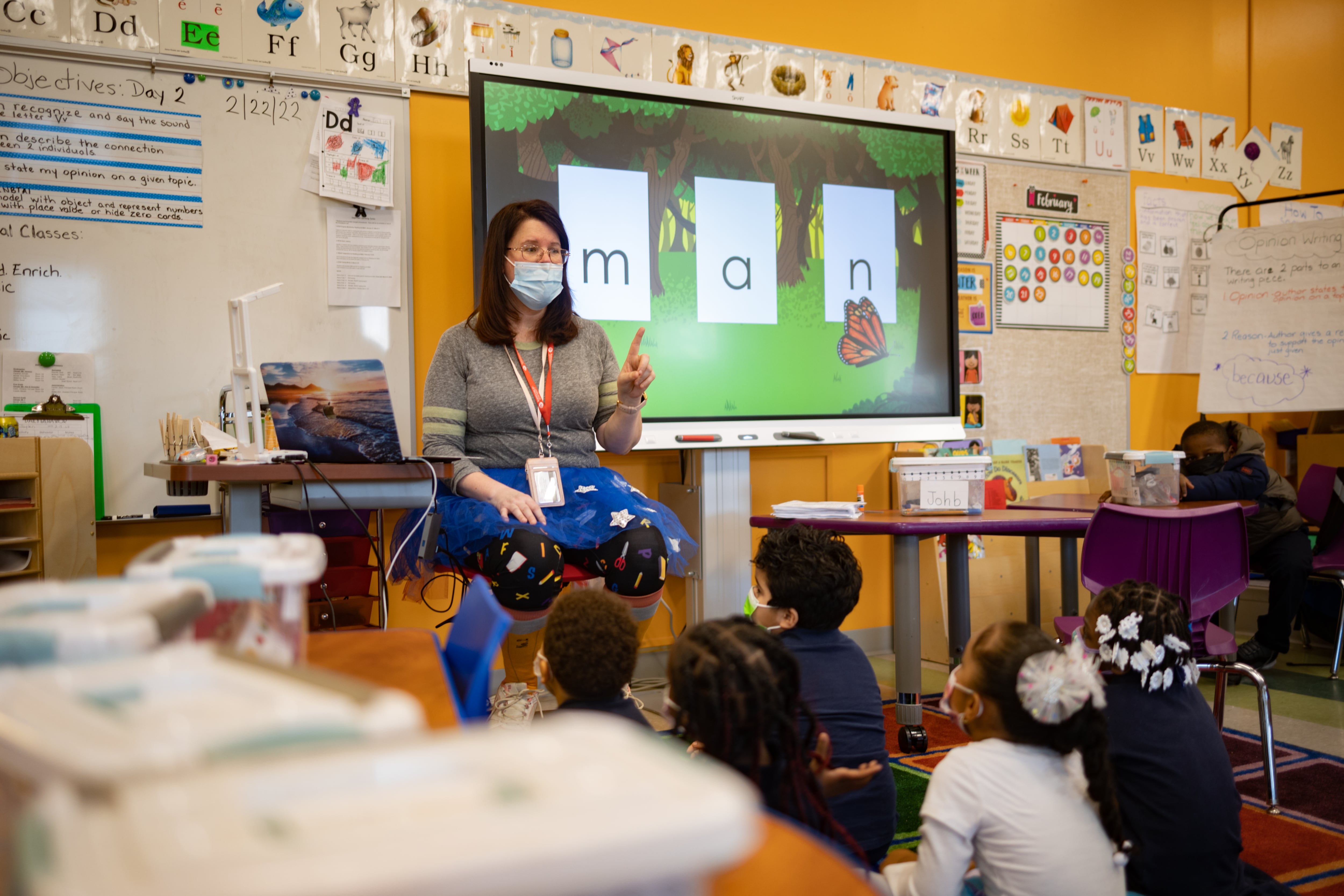 A teacher sits in front of her class with mask on.