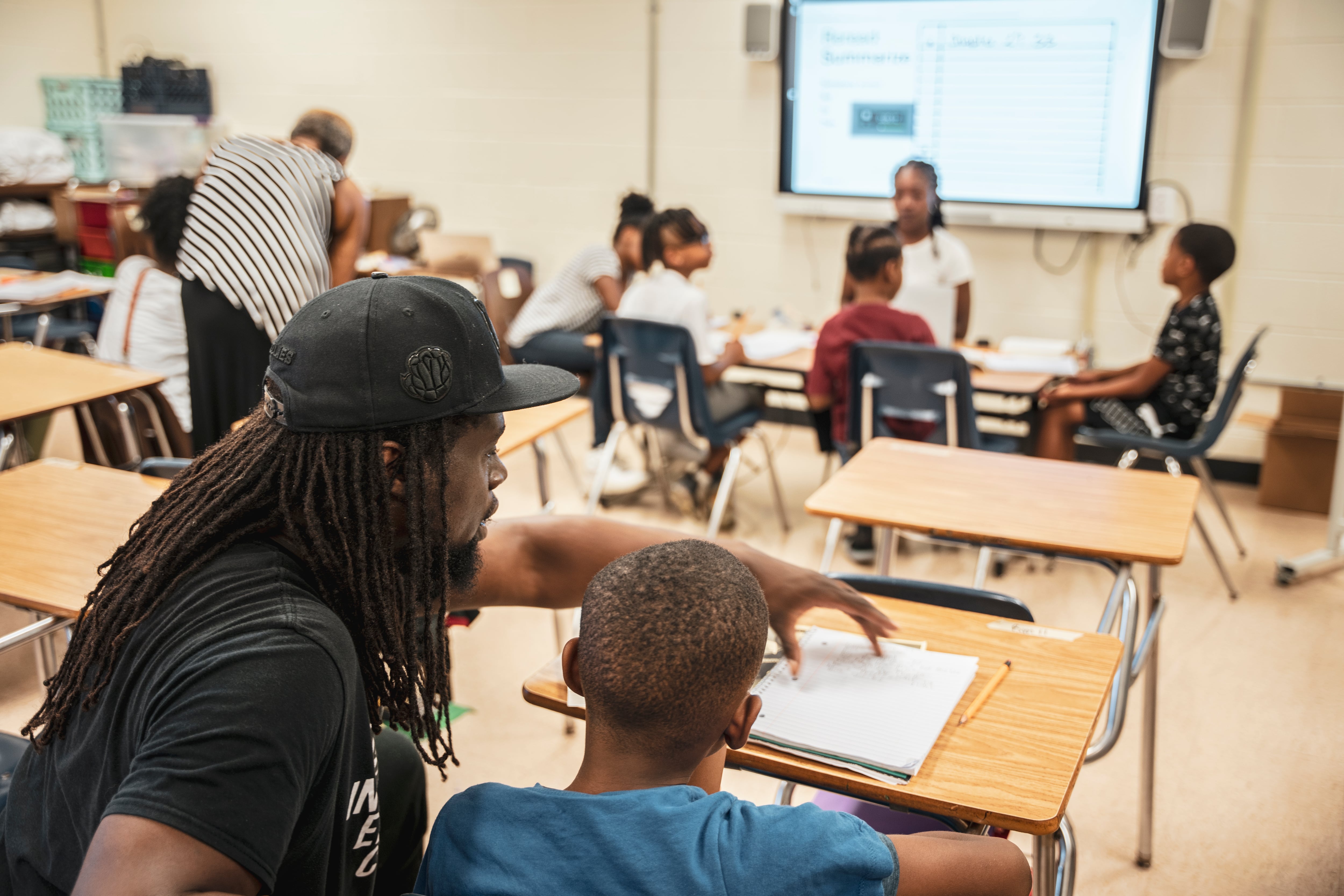 A man in a black hat and a black shirt touches paper on a desk as a boy in a blue shirt looks on.