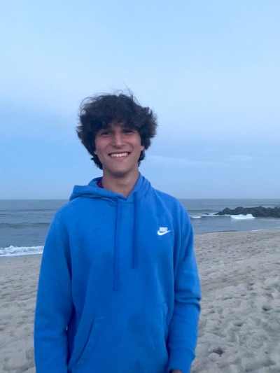 A high school boy wearing a blue hoodie smiles at the camera with the beach and ocean in the background.