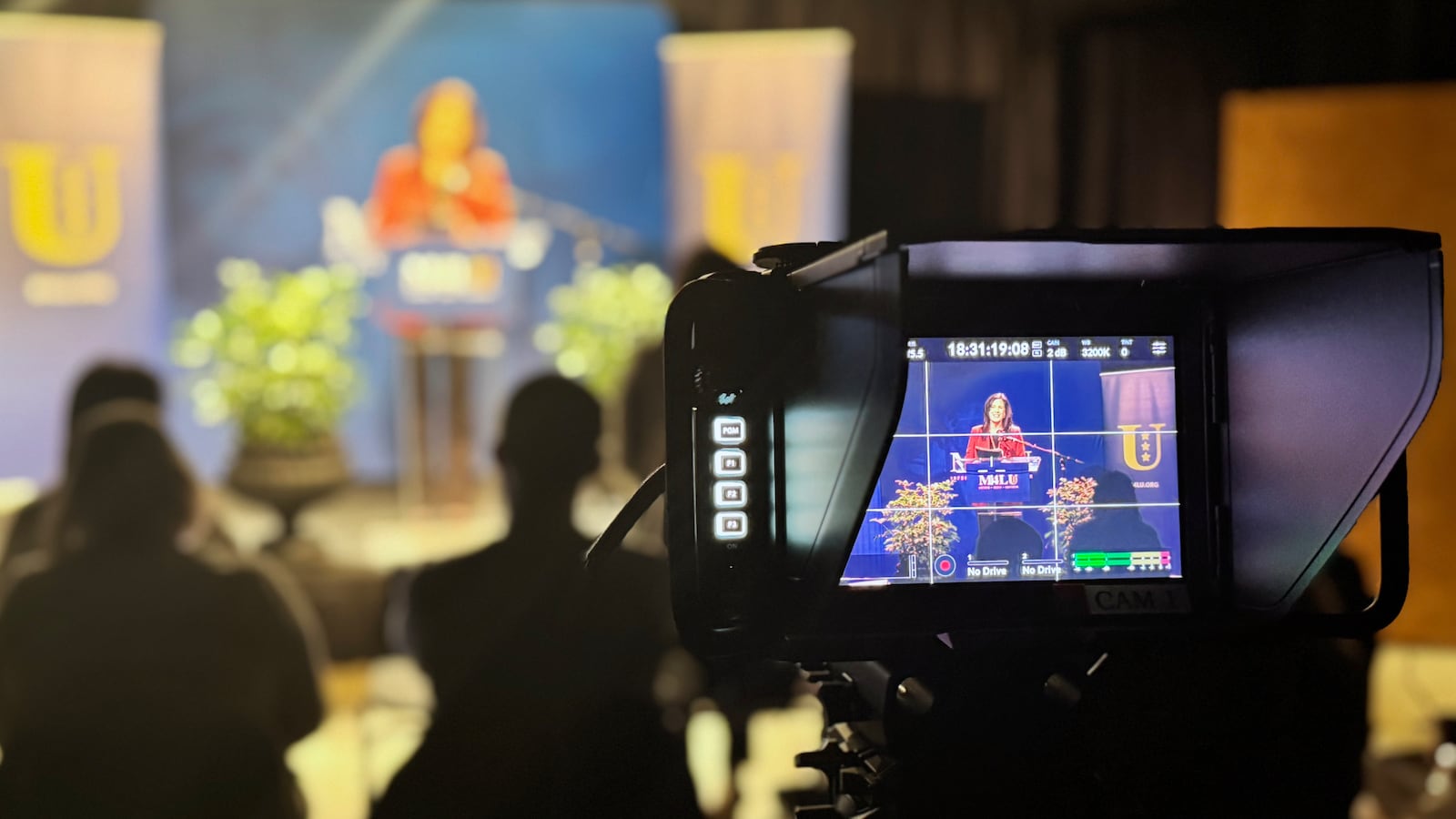 A view from behind a large camera viewfinder in focus on a woman speaking behind a podium and in front of a blue background while in the background of the viewfinder is a blurry scene.