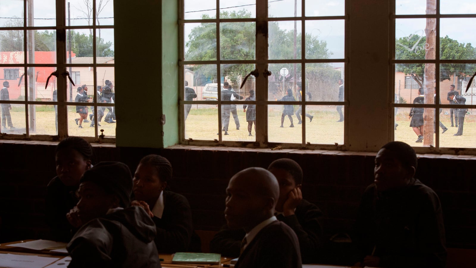Students sit in a dark classroom at their desks in South Africa, as other children play in a field outside.