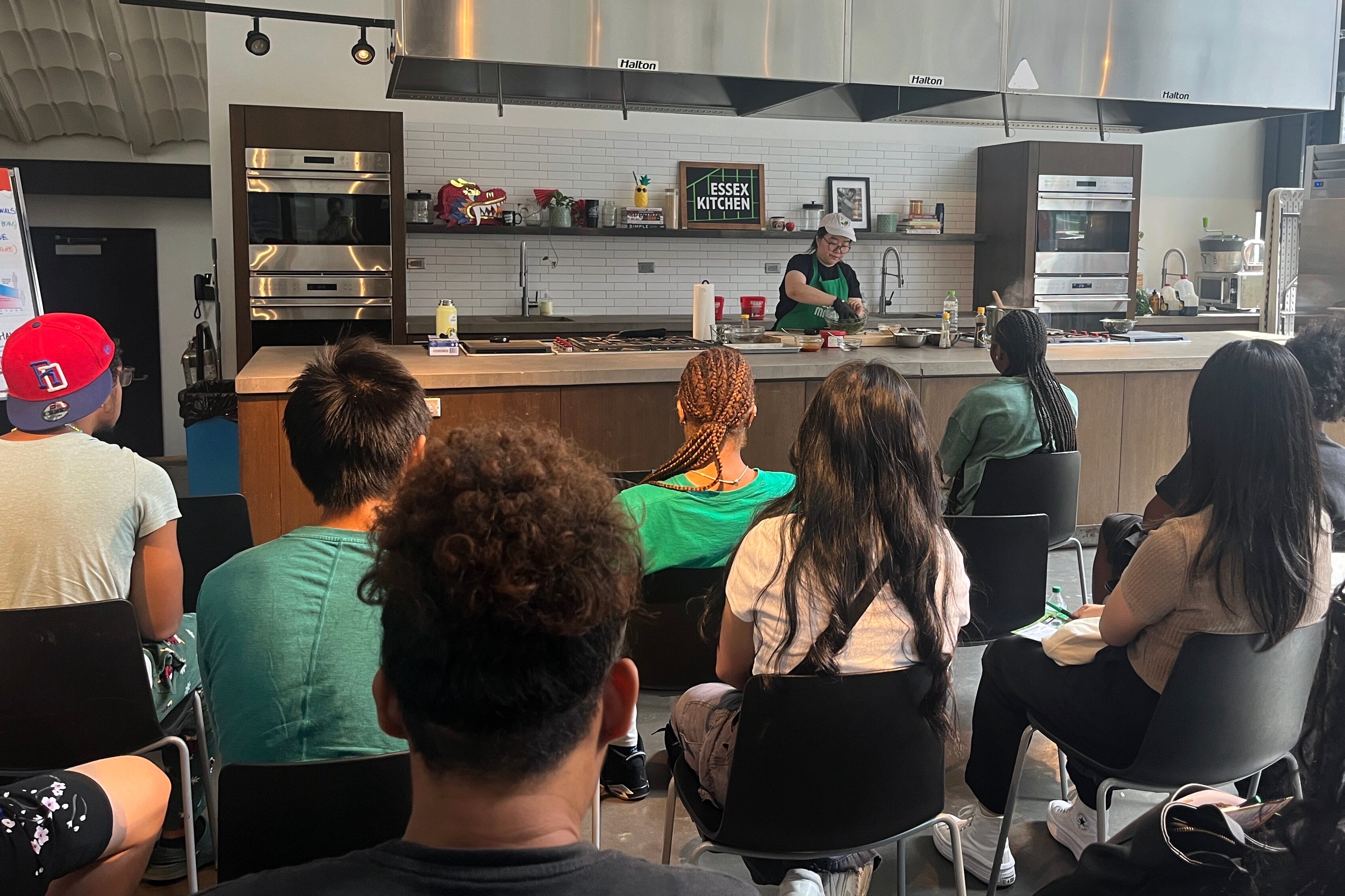 Students sit in chairs inside of an industrial kitchen while one person works behind the counter cooking.