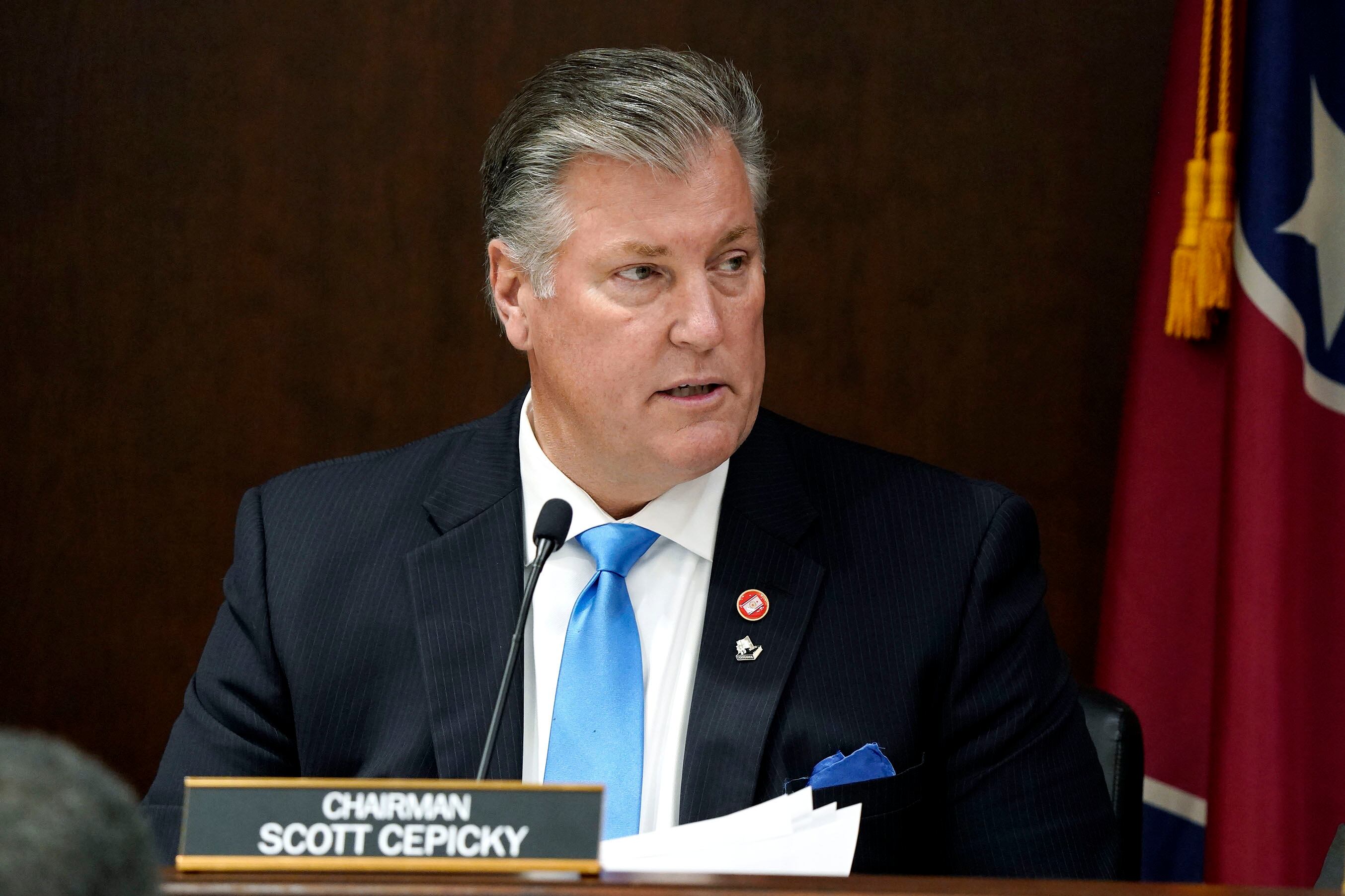 A man wearing a suit and blue necktie speaks into a microphone in a legislative chamber with the Tennessee state flag in the background.