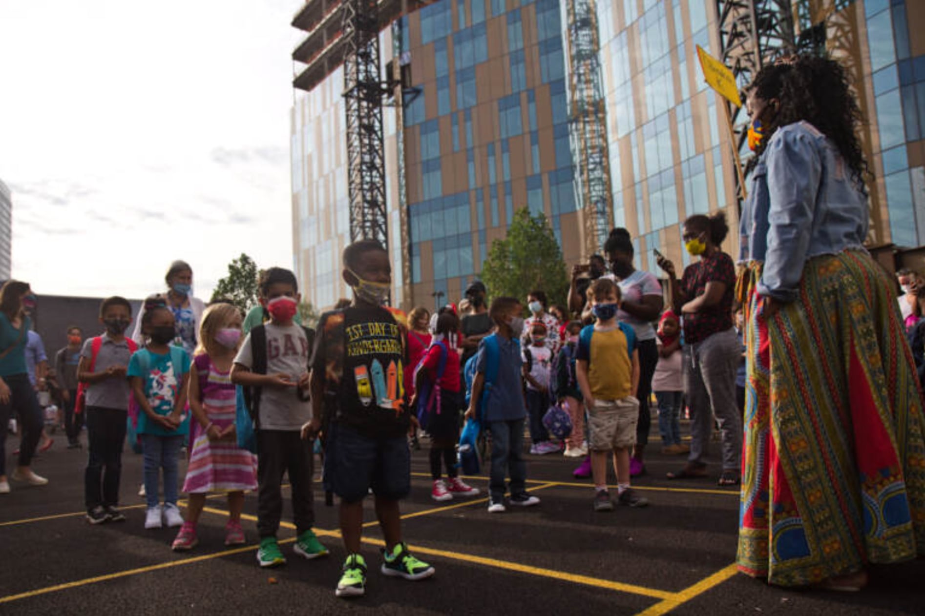 Philadelphia students, wearing protective masks, arrive for the first day of classes and stand around yellow lines on the ground. A building is in the background.