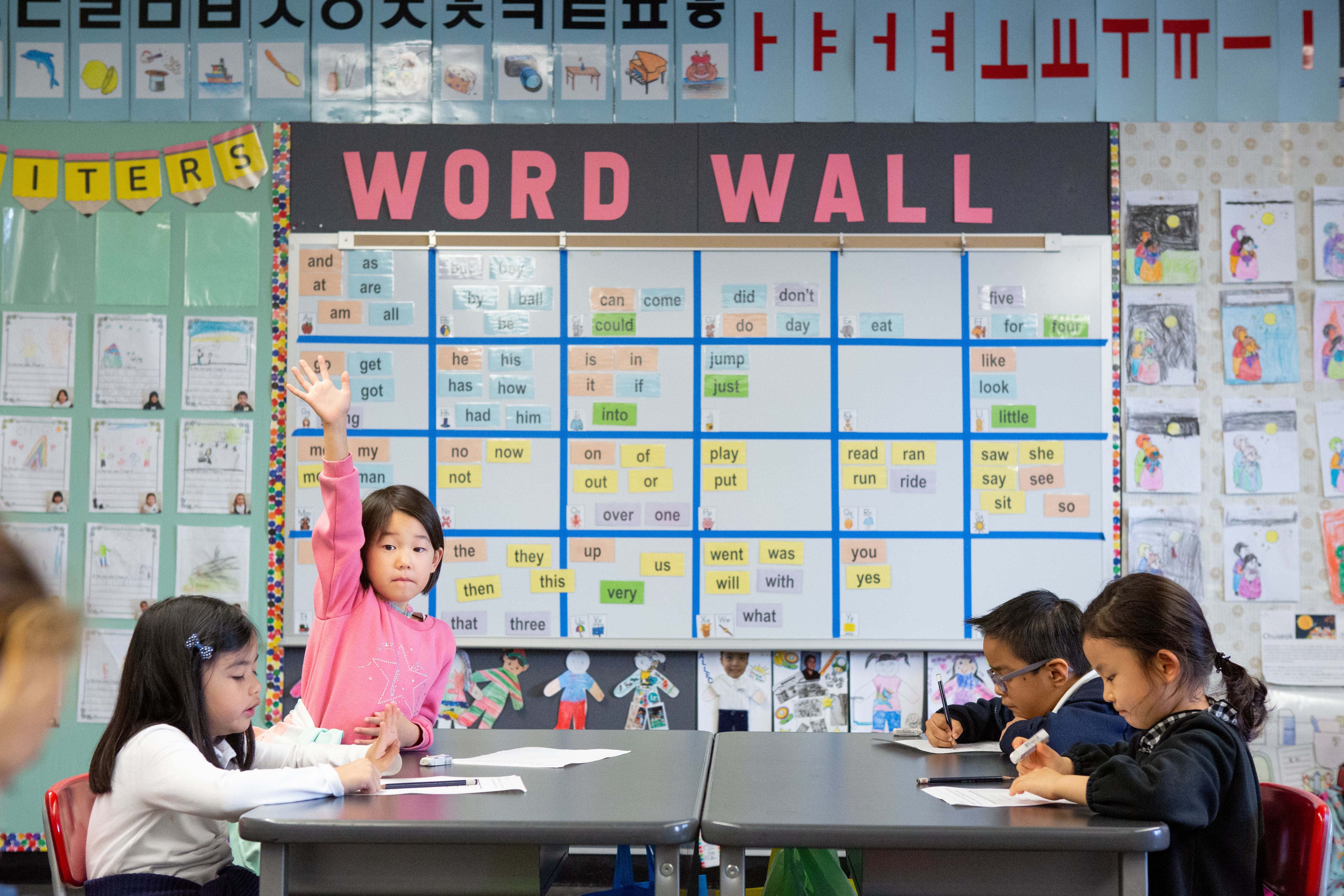 Four young kindergarten students sitting at desks in front of a colorful wall. One is raising her hand and looking at the camera.