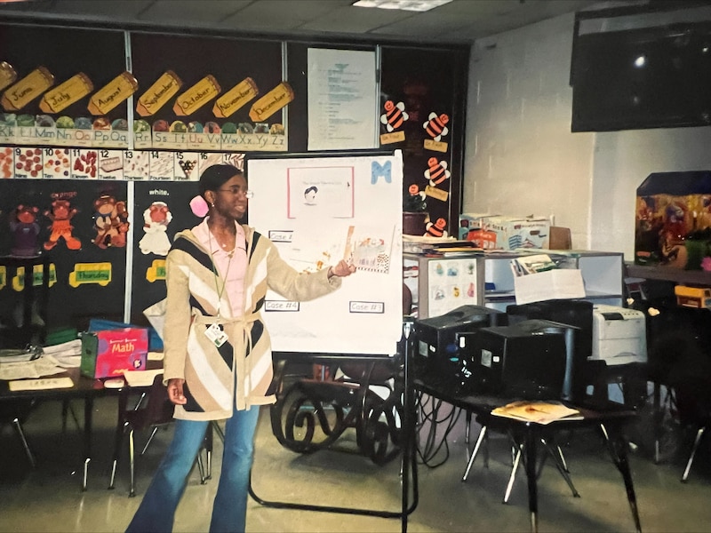 A photograph of a young Black woman teaching in a classroom.