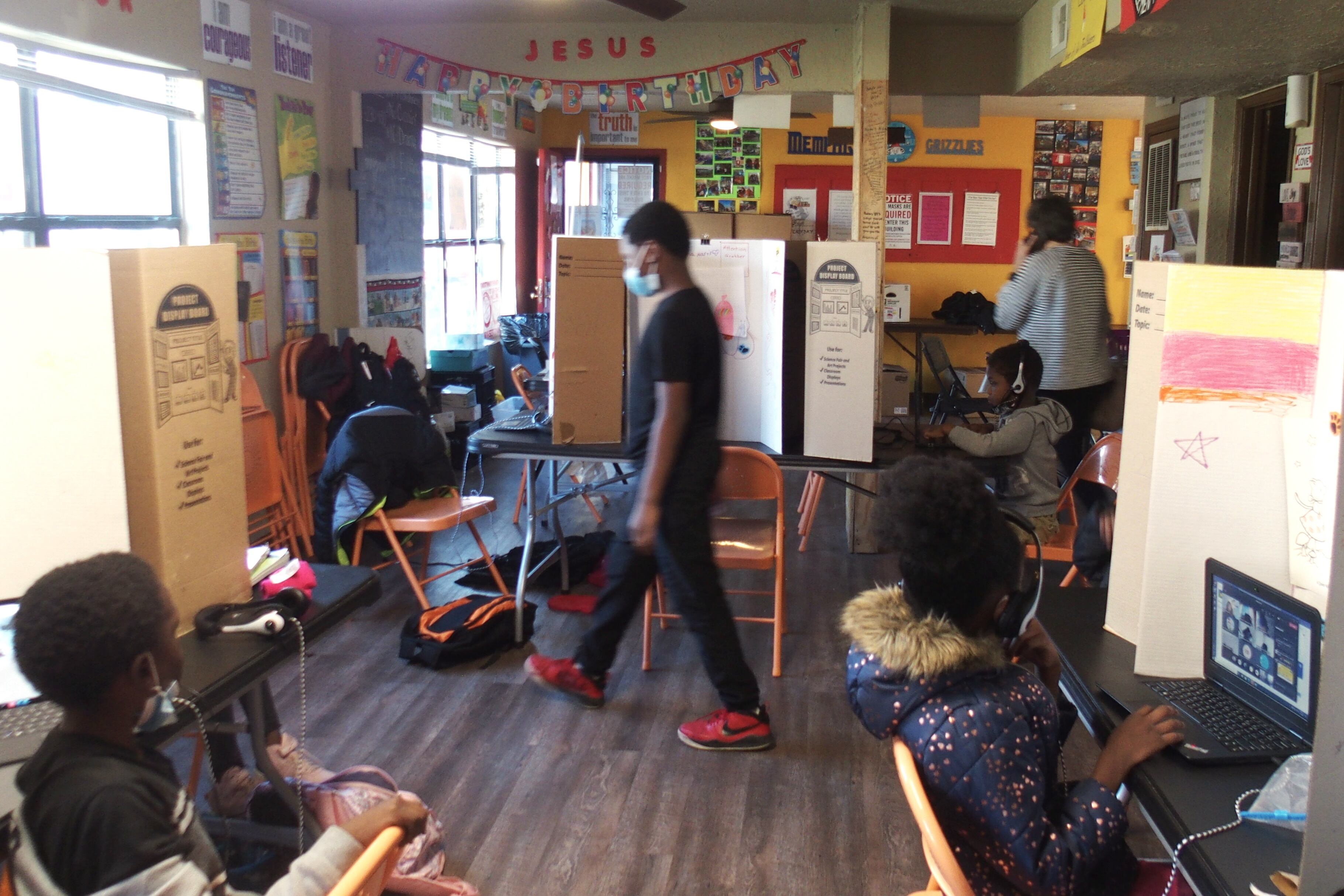 A student in a mask walks back to his seat at a table with cardboard dividers as students attend online classes via laptops