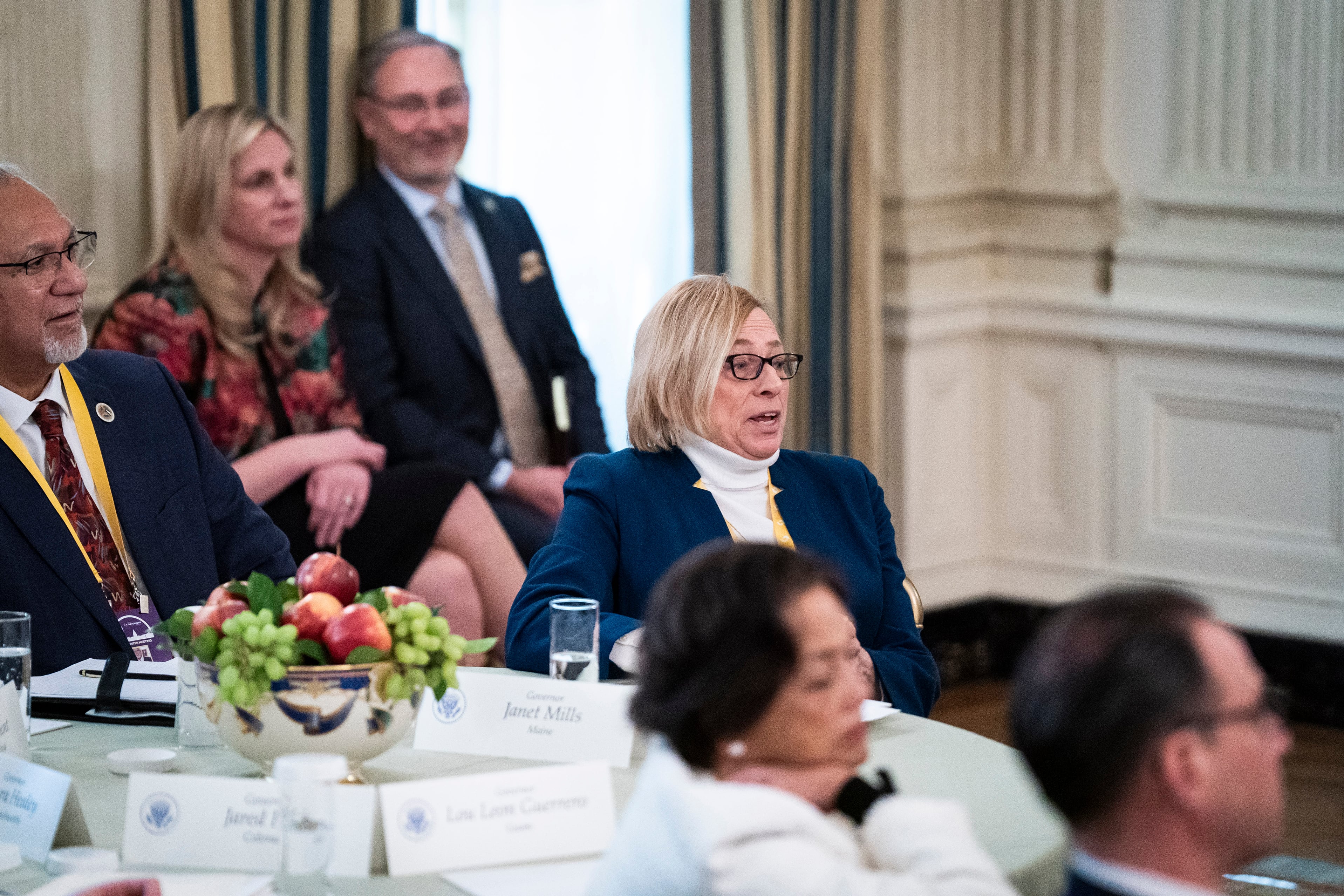 A white woman wearing a blue suit jacket and short blonde hair sits at a round table in a room full of other people wearing business clothes.