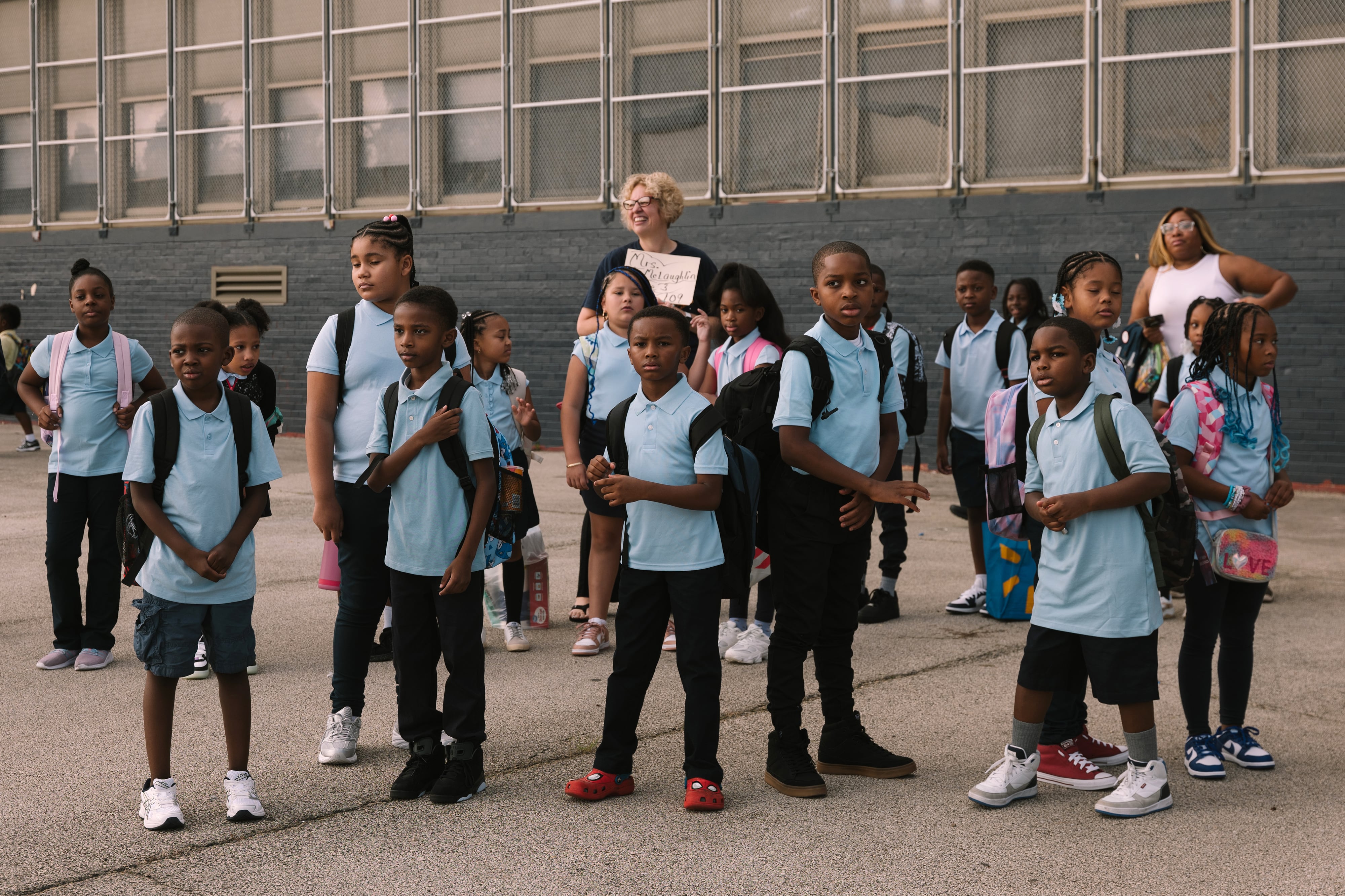 A group of elementary students in blue uniforms and black pants stand outside of a large building on the first day of school.